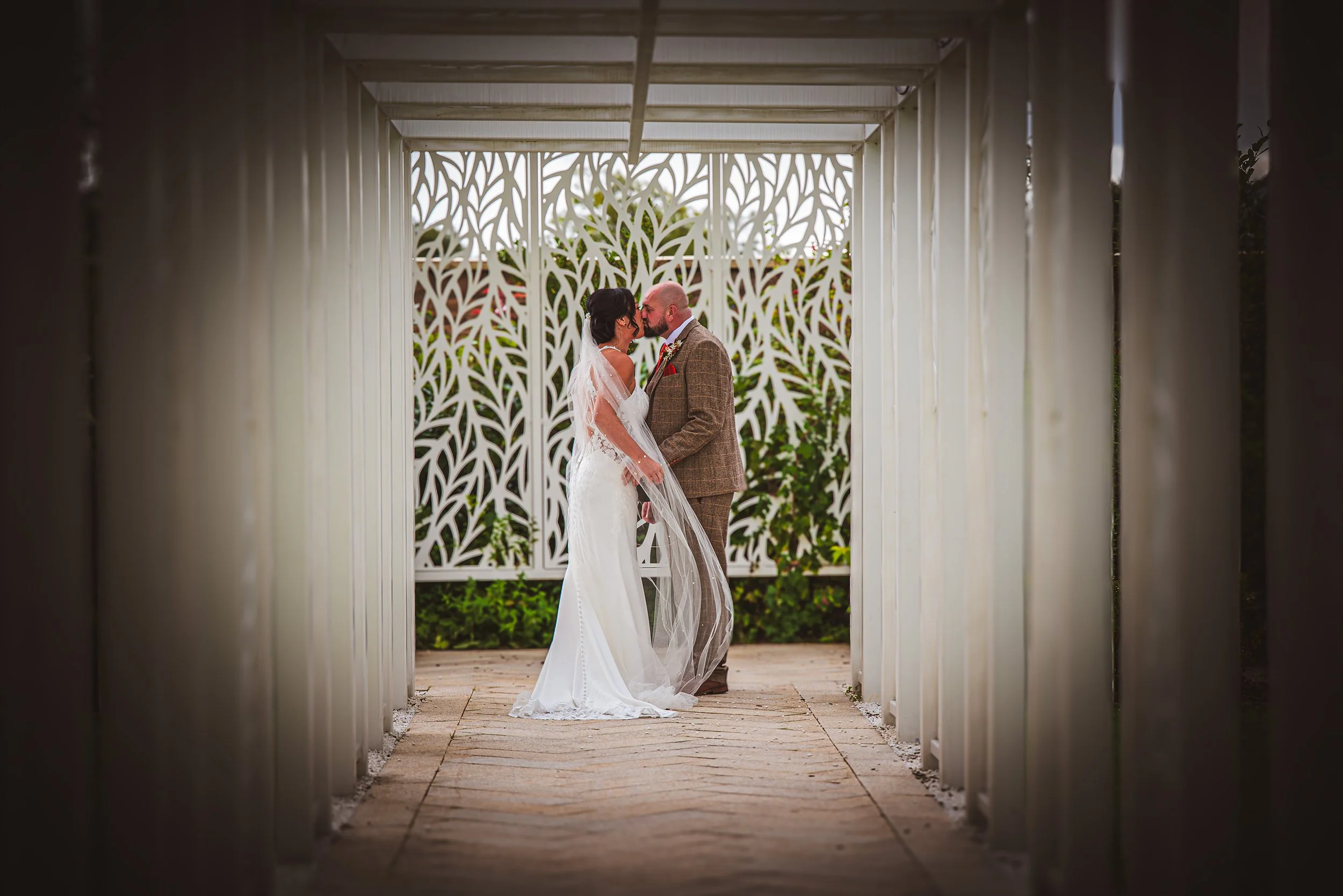 Bride and groom share a kiss in a decorative outdoor structure with intricate white lattice panels and lush greenery in the background.