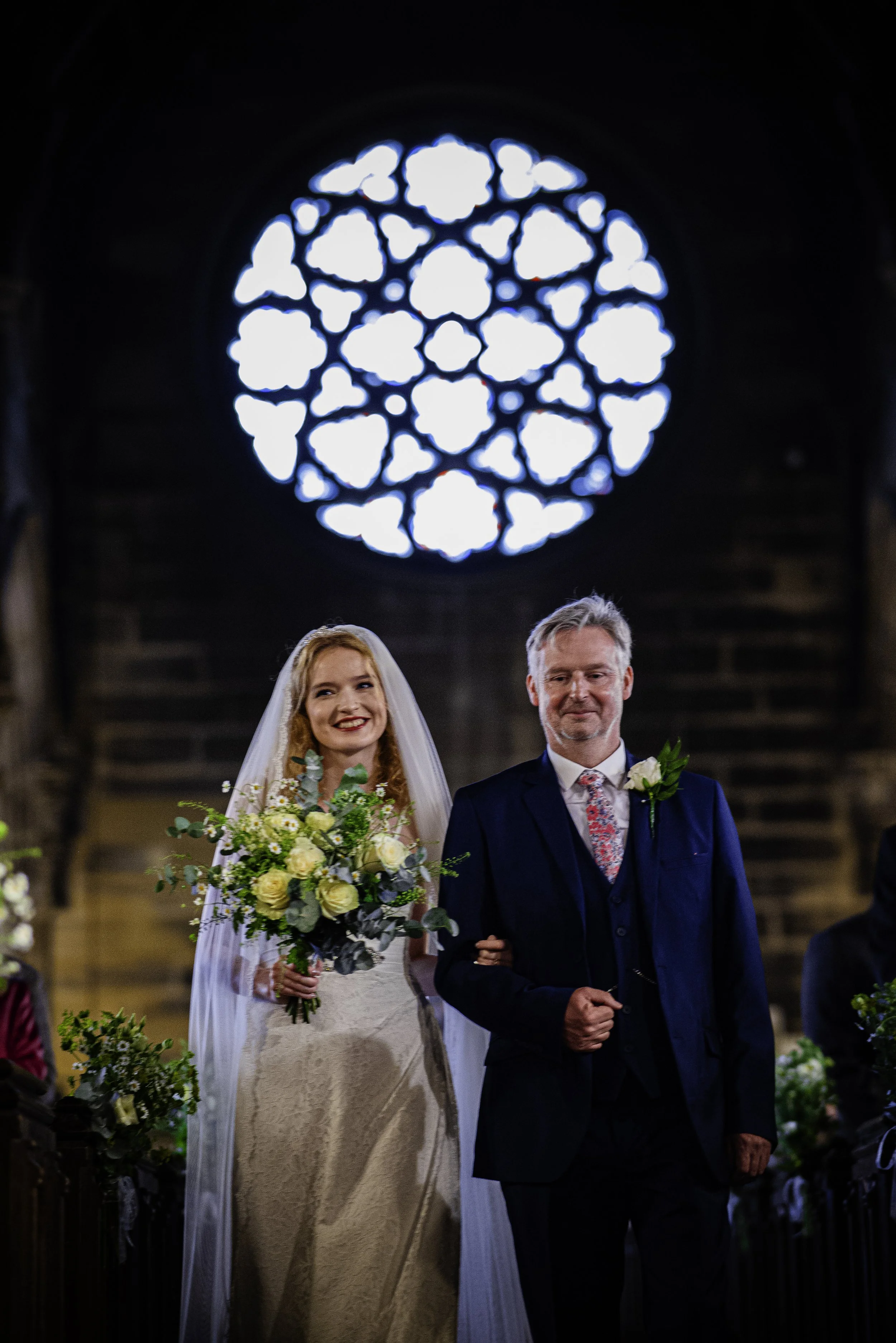 A bride in a wedding dress holding a bouquet of white and yellow roses walks down the aisle with a groom in a navy suit, inside a church with stained glass window behind them.