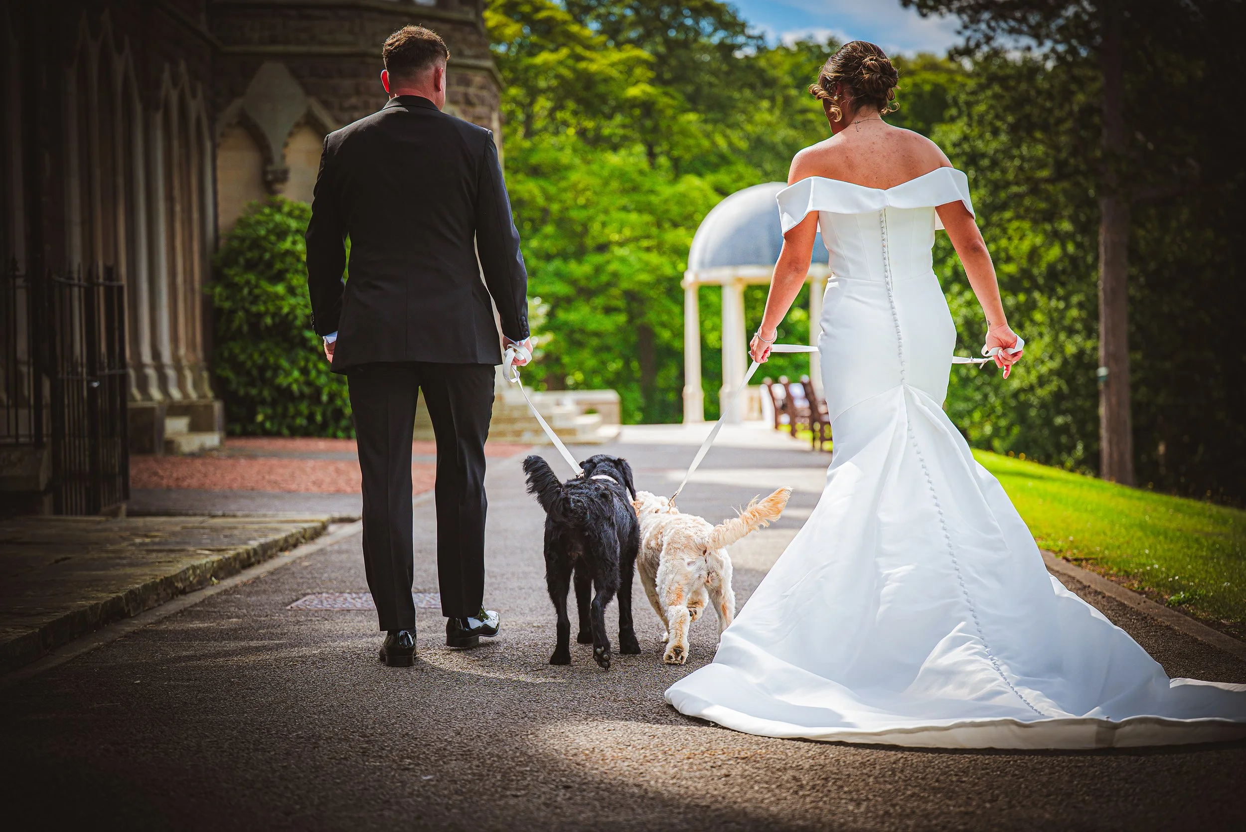Bride and groom walking away together on a wedding day, each holding a leash attached to their two dogs, in an outdoor setting with lush green trees and a structure with a domed roof in the background.