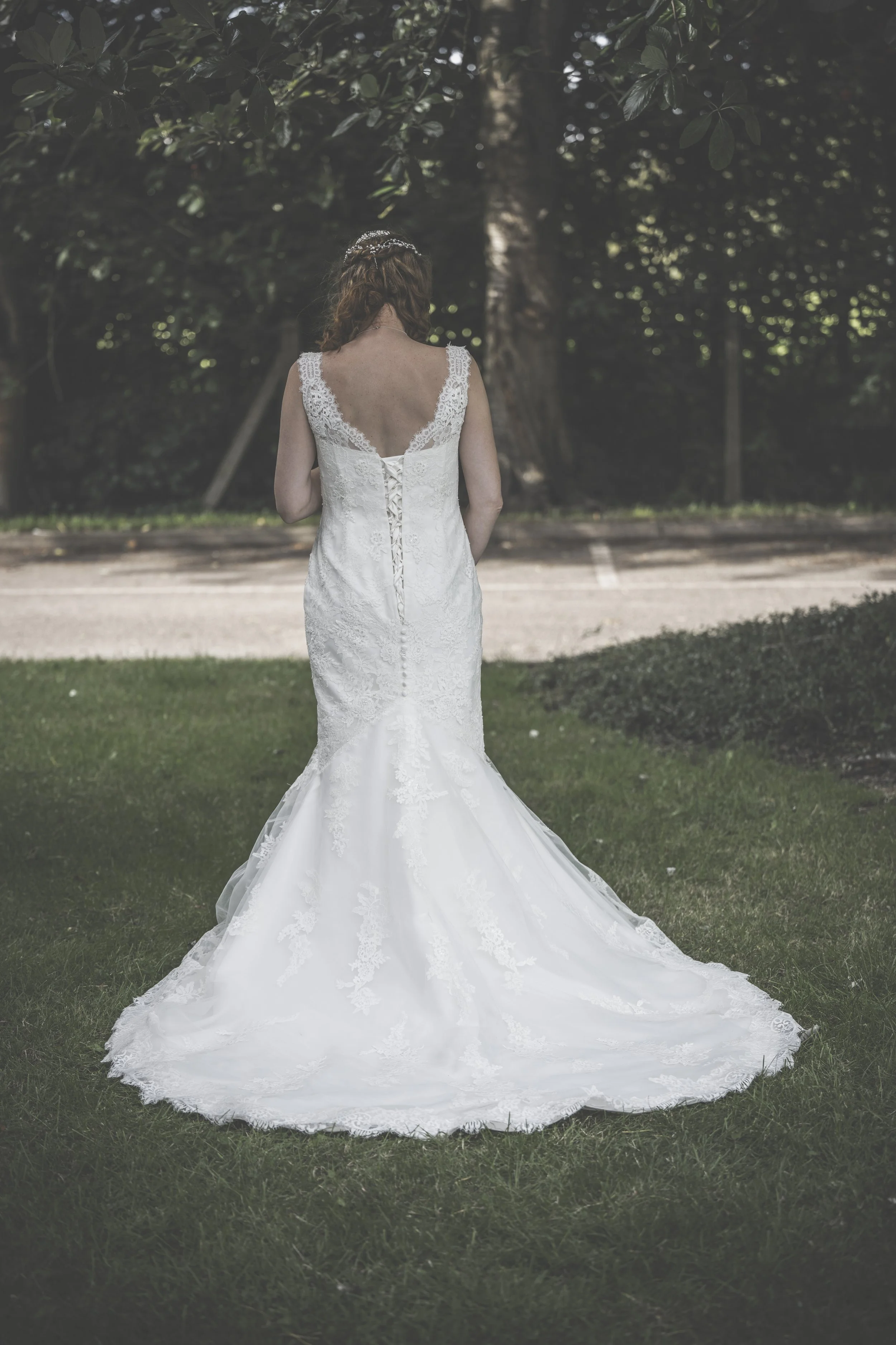 A bride in a white gown with lace details, standing outdoors on grass with trees in the background, facing away from the camera.