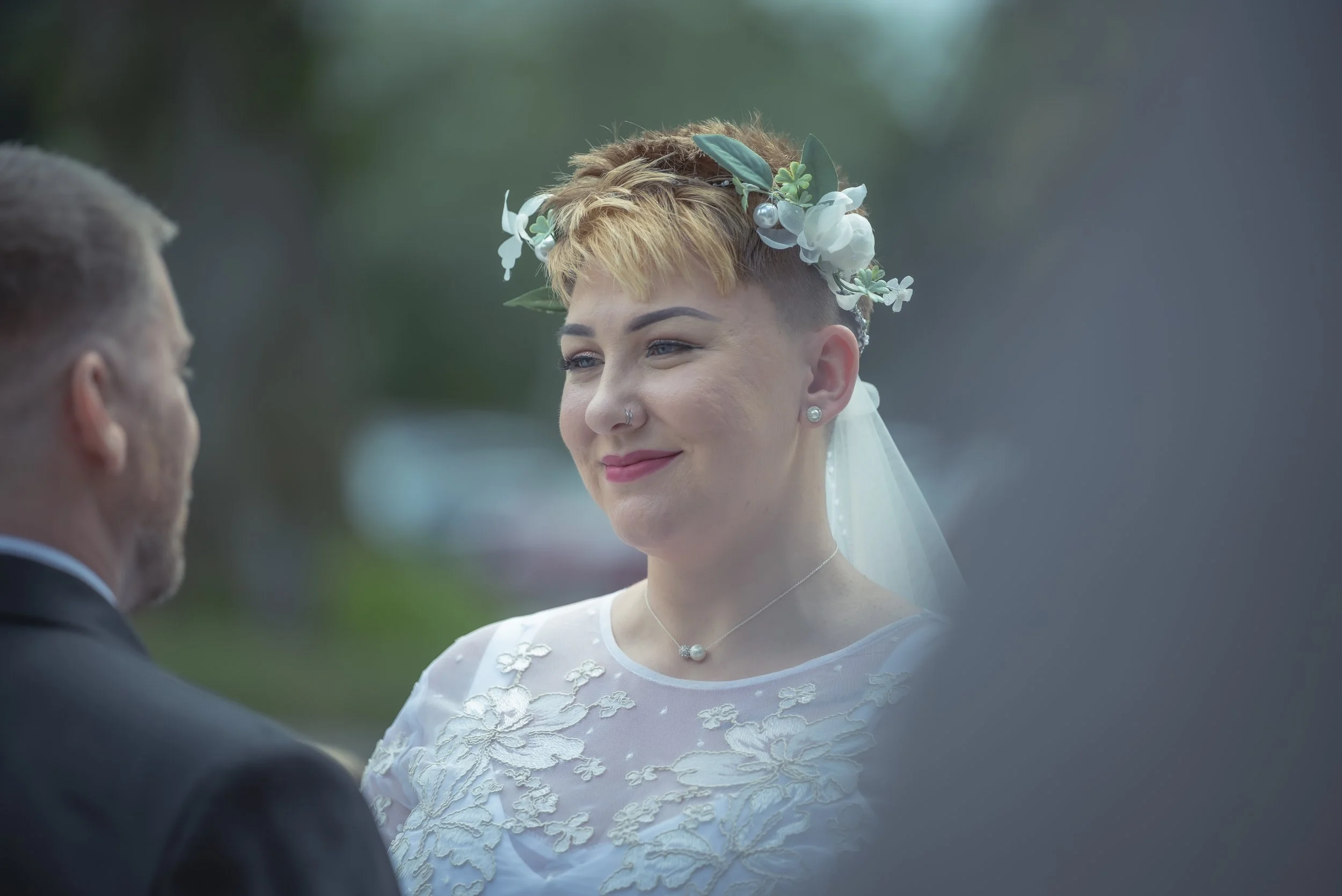 Bride with short blonde hair, floral headband, white embroidered dress, and pearl jewelry, smiling during wedding ceremony.