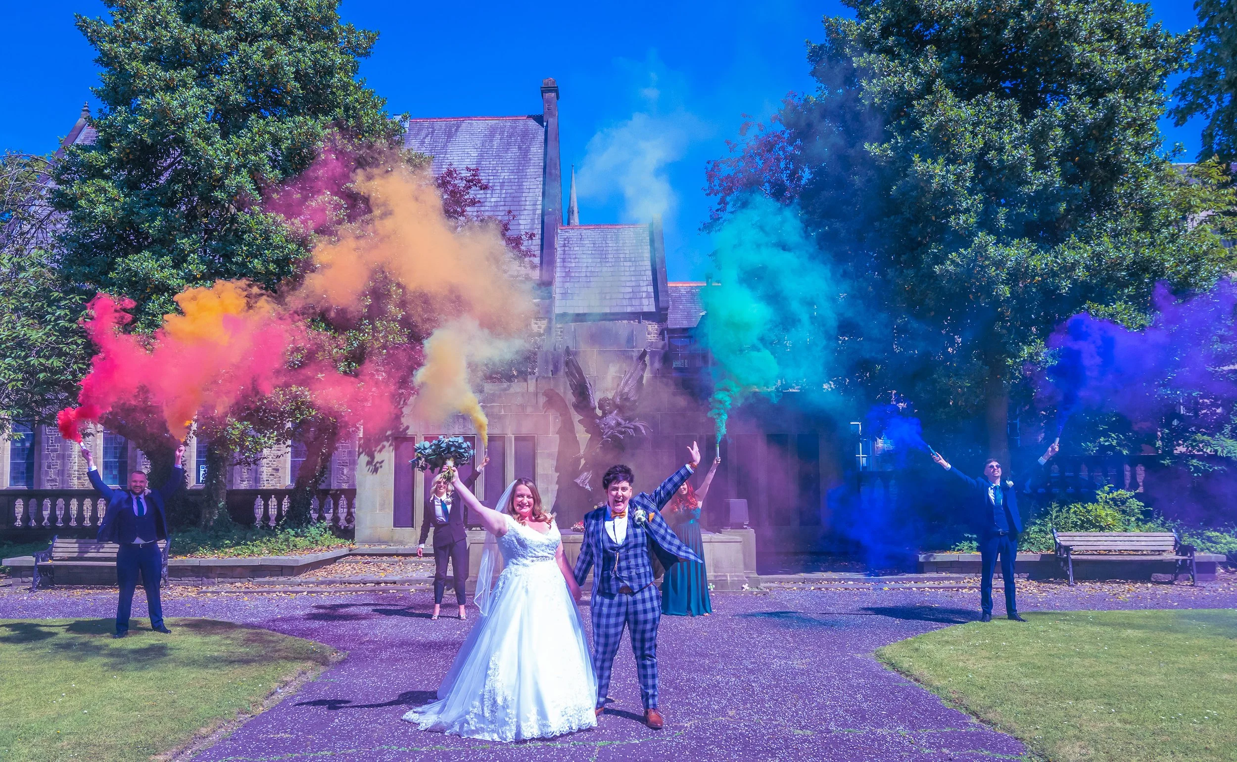 Wedding celebration with couple in the foreground, smiling and holding smoke bombs emitting colorful smoke, surrounded by friends in formal attire in front of a historic building with statue and trees.