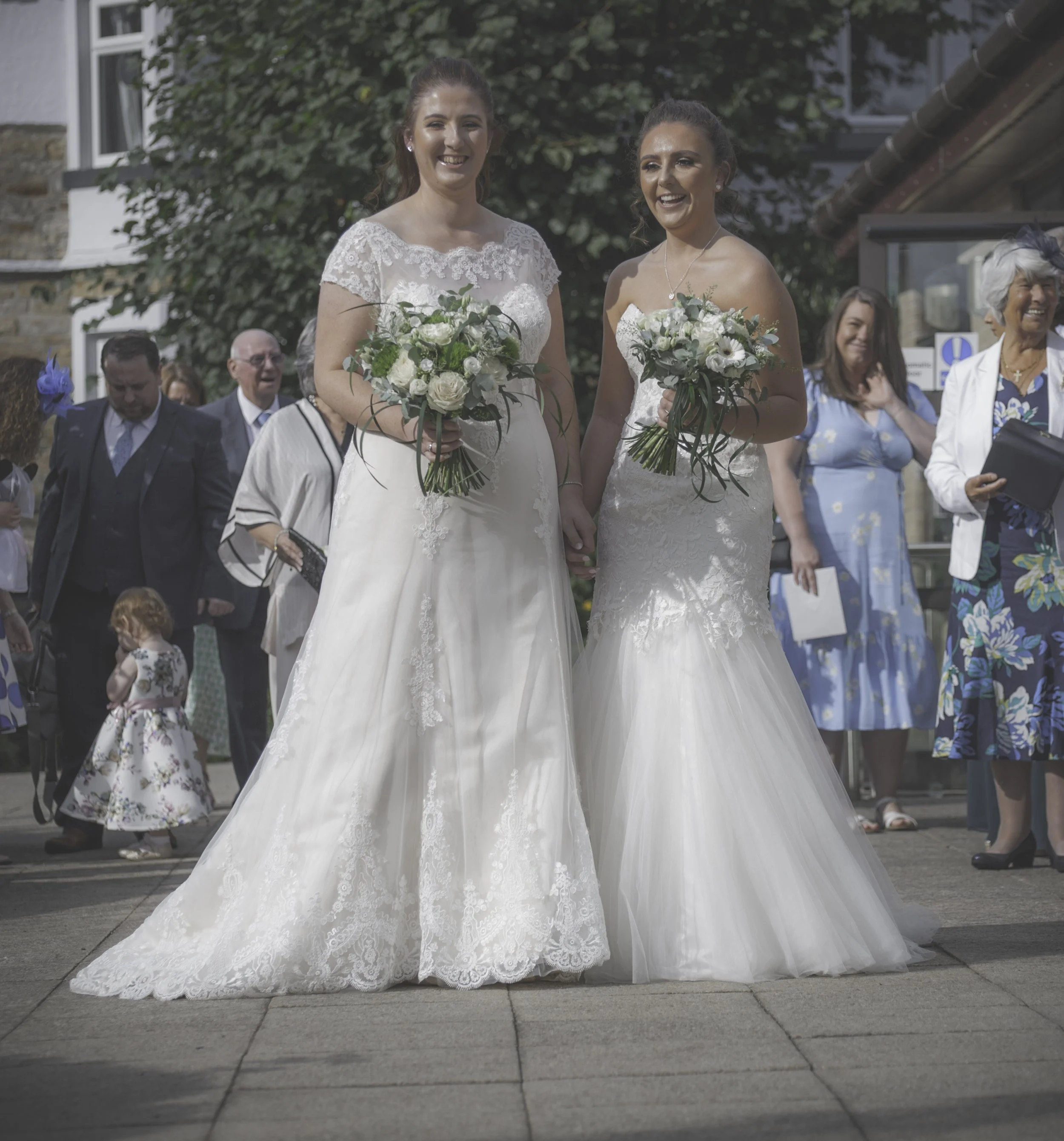 Two brides holding hands, both in white wedding gowns and holding bouquets, smiling outdoors with guests in the background.