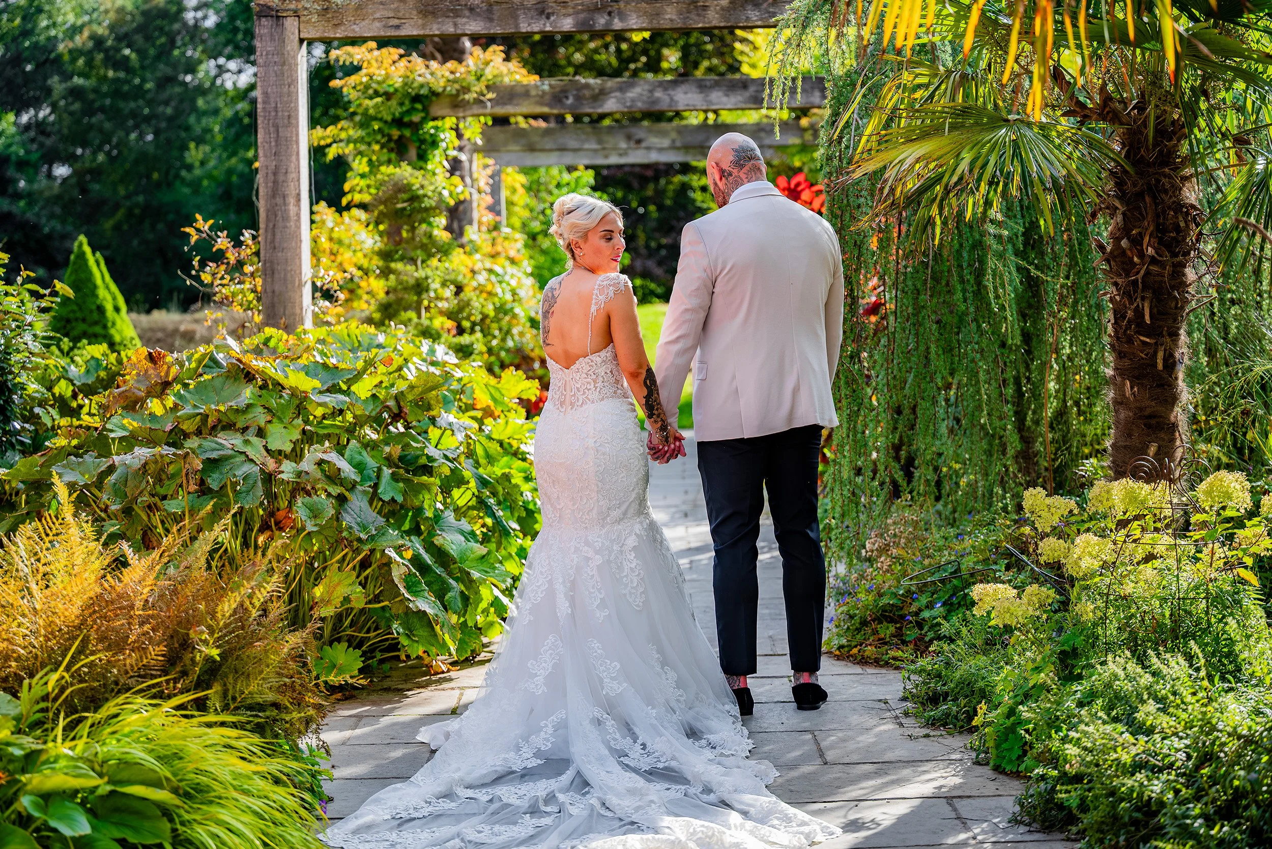 A bride and groom walking hand in hand in a lush garden setting, viewed from behind, with the bride in a white lace wedding gown and the groom in a light-colored jacket and dark pants.