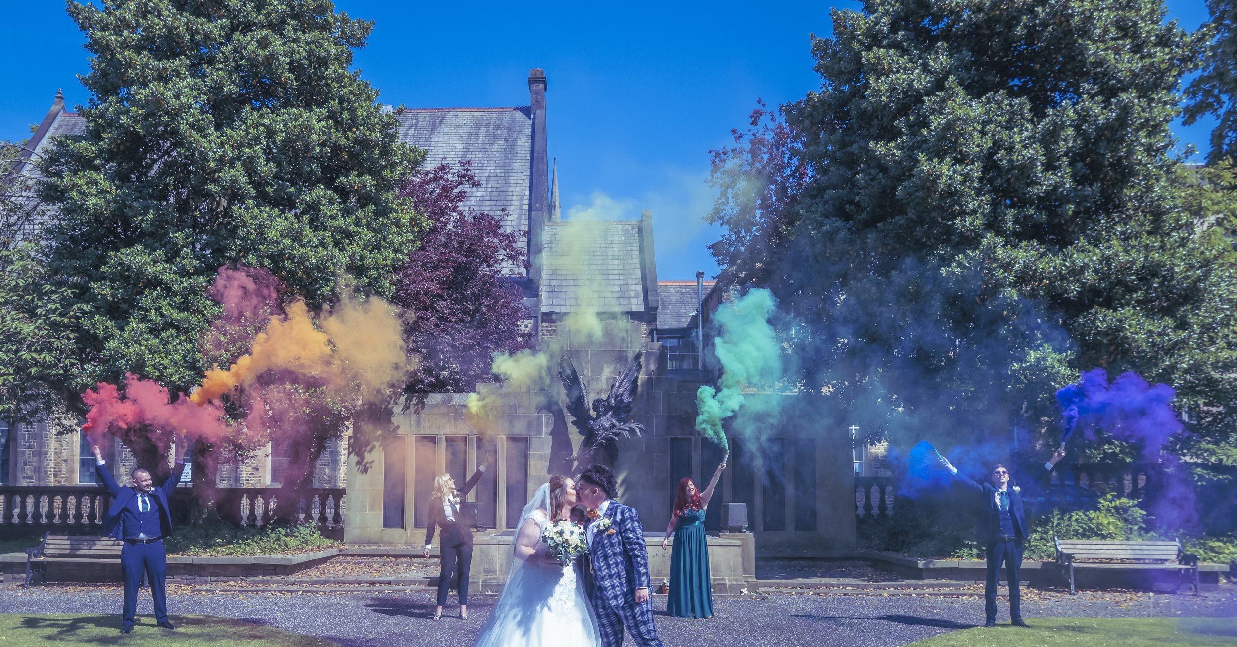 A wedding celebration outdoors with a bride and groom kissing, surrounded by friends holding colored smoke bombs in blue, purple, yellow, green, orange, and pink, in front of a stone building with large trees and a bright blue sky.