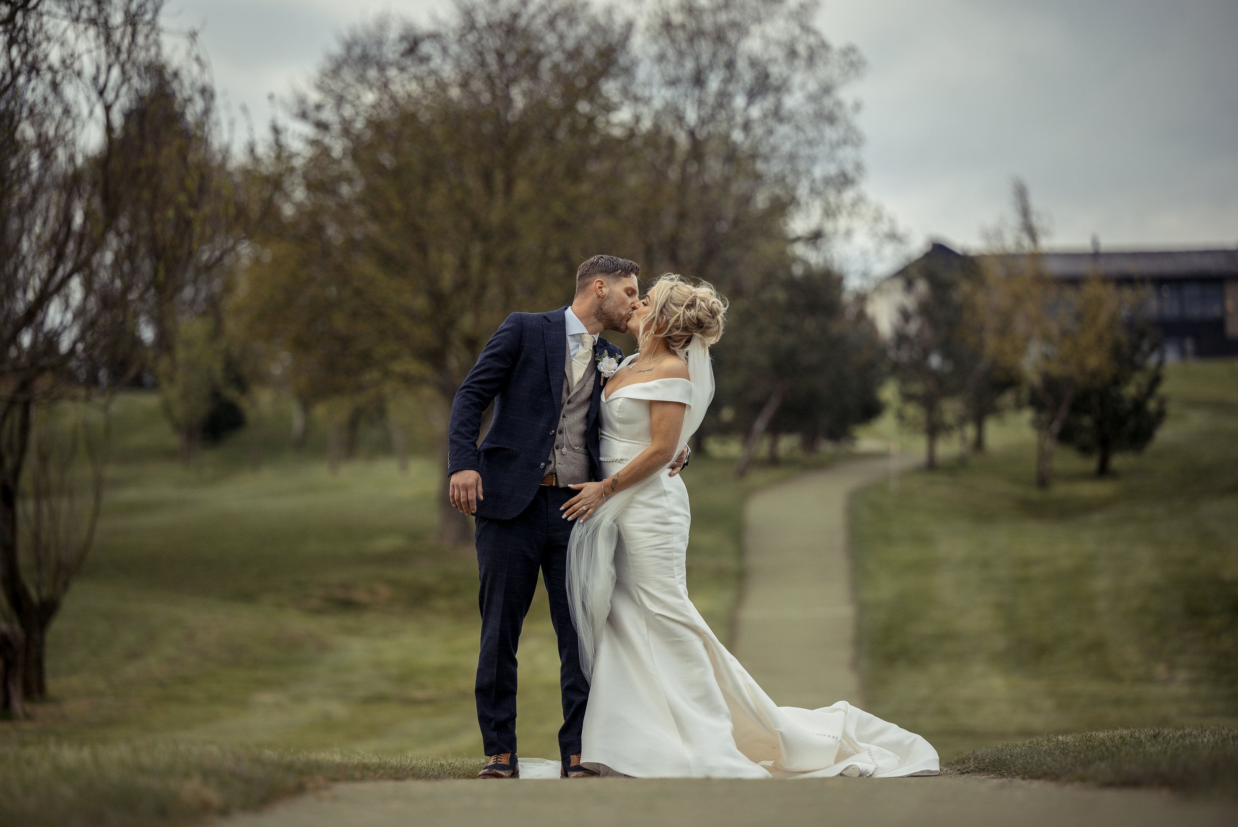 A bride and groom share a kiss outdoors on a path surrounded by trees during a wedding photoshoot.