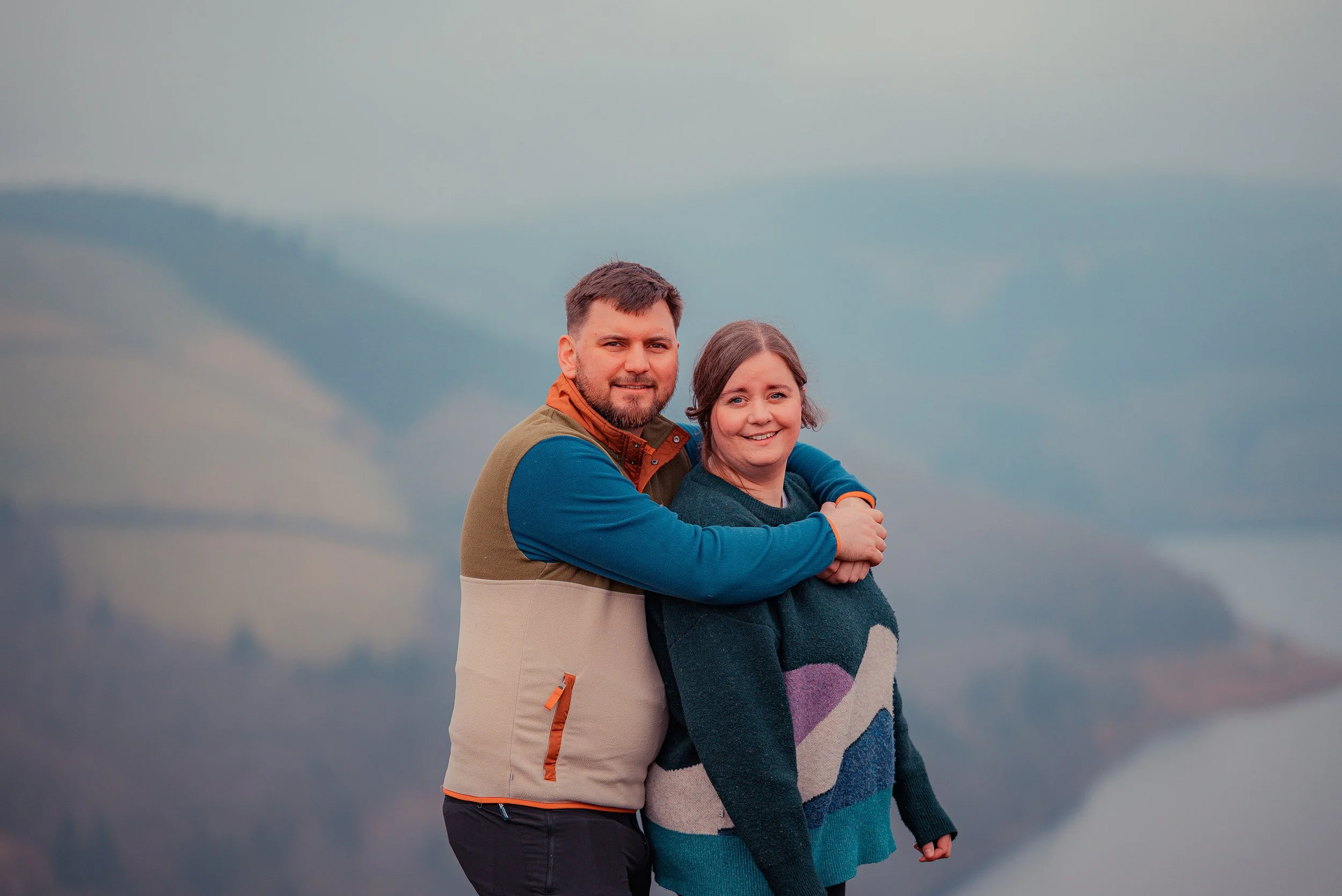 A man and woman standing together outdoors with a scenic, foggy mountain and water in the background. The man has his arms around the woman, both smiling at the camera.