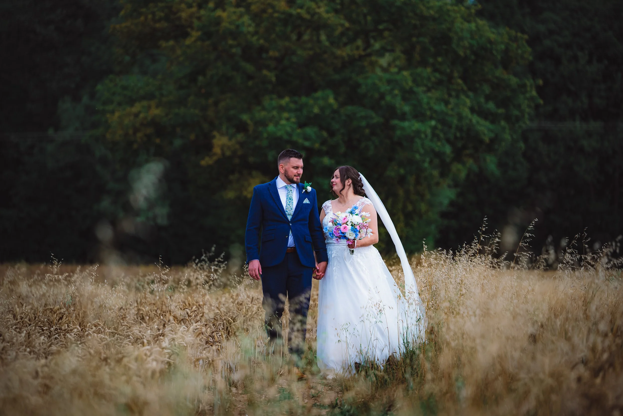 A bride and groom standing hand in hand in a field of tall, golden grass with a dense green forest in the background. The bride is wearing a white wedding dress with a long veil and holding a colorful bouquet. The groom is dressed in a navy blue suit
