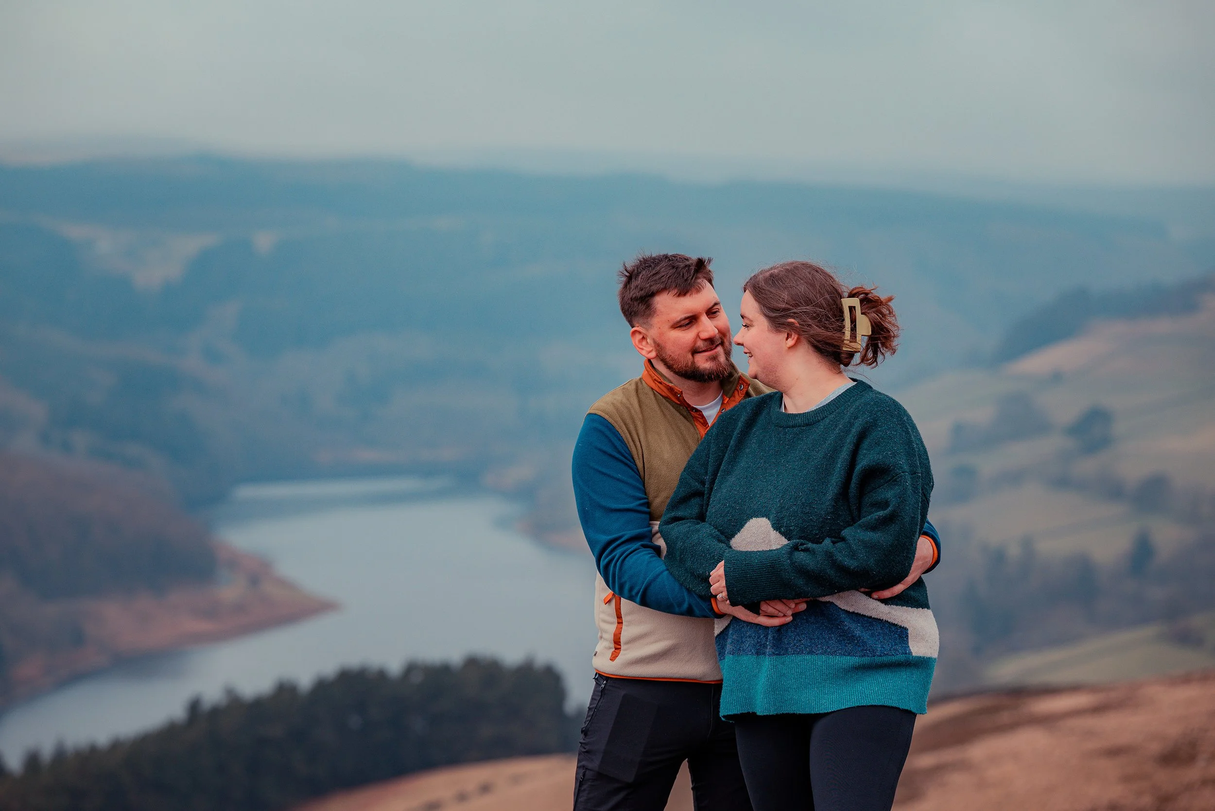 A couple embracing outdoors with a valley and lake in the background.