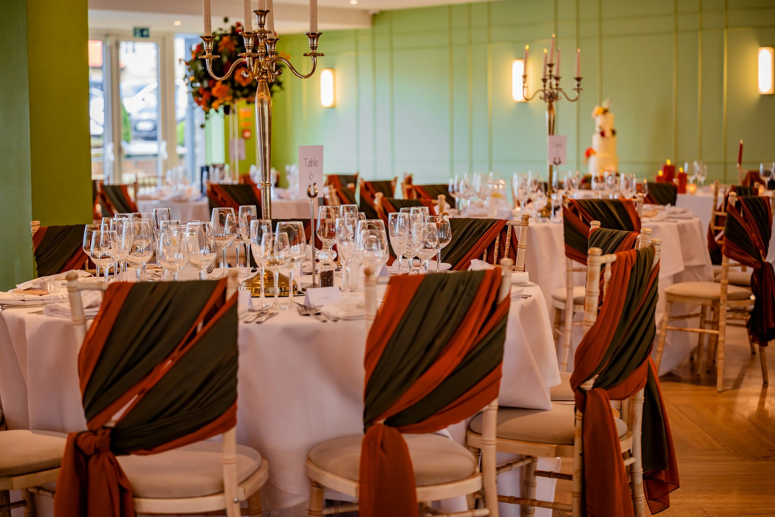 Elegant banquet hall decorated for a special event, with round tables dressed in white tablecloths, set with multiple wine glasses, and chairs draped with black and rust-colored fabric. Tall candelabras and floral centerpieces adorn the tables. Soft 