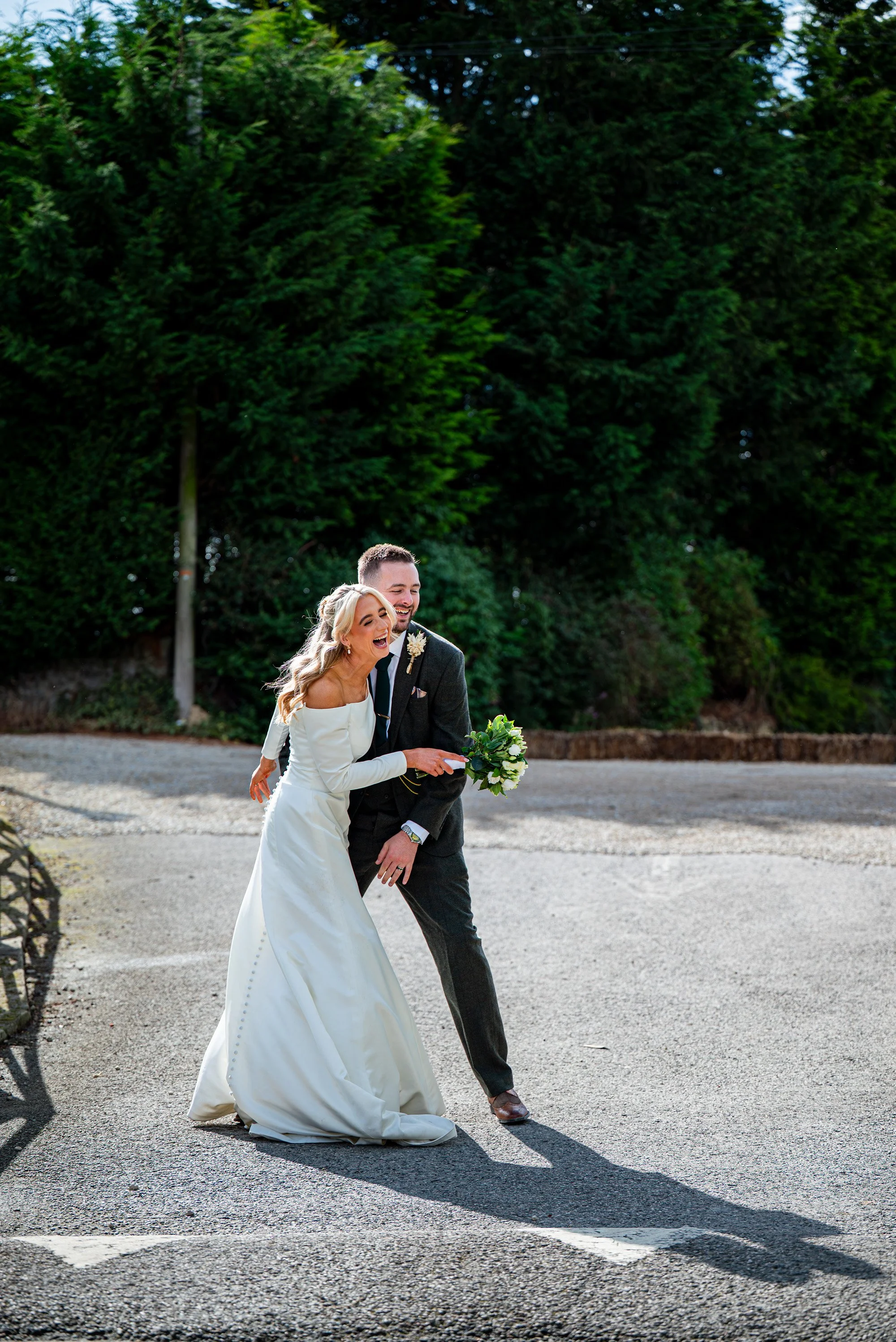 A bride and groom sharing a joyful moment outdoors, standing on a paved area with lush green trees in the background. The bride is wearing a white wedding gown, and the groom is in a dark suit holding a bouquet of white flowers.