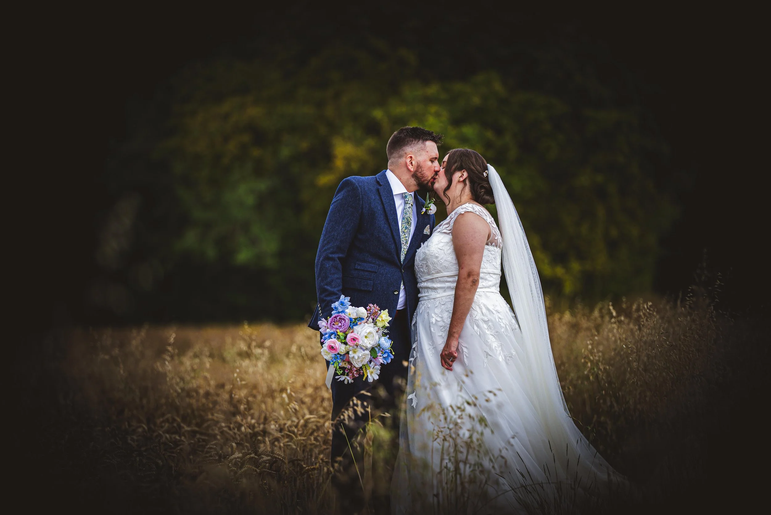A newlywed couple kissing outdoors in a field with tall grass, a man in a blue suit holding a colorful bouquet, and a woman in a white wedding dress with a veil, surrounded by a blurred green and yellow foliage background.