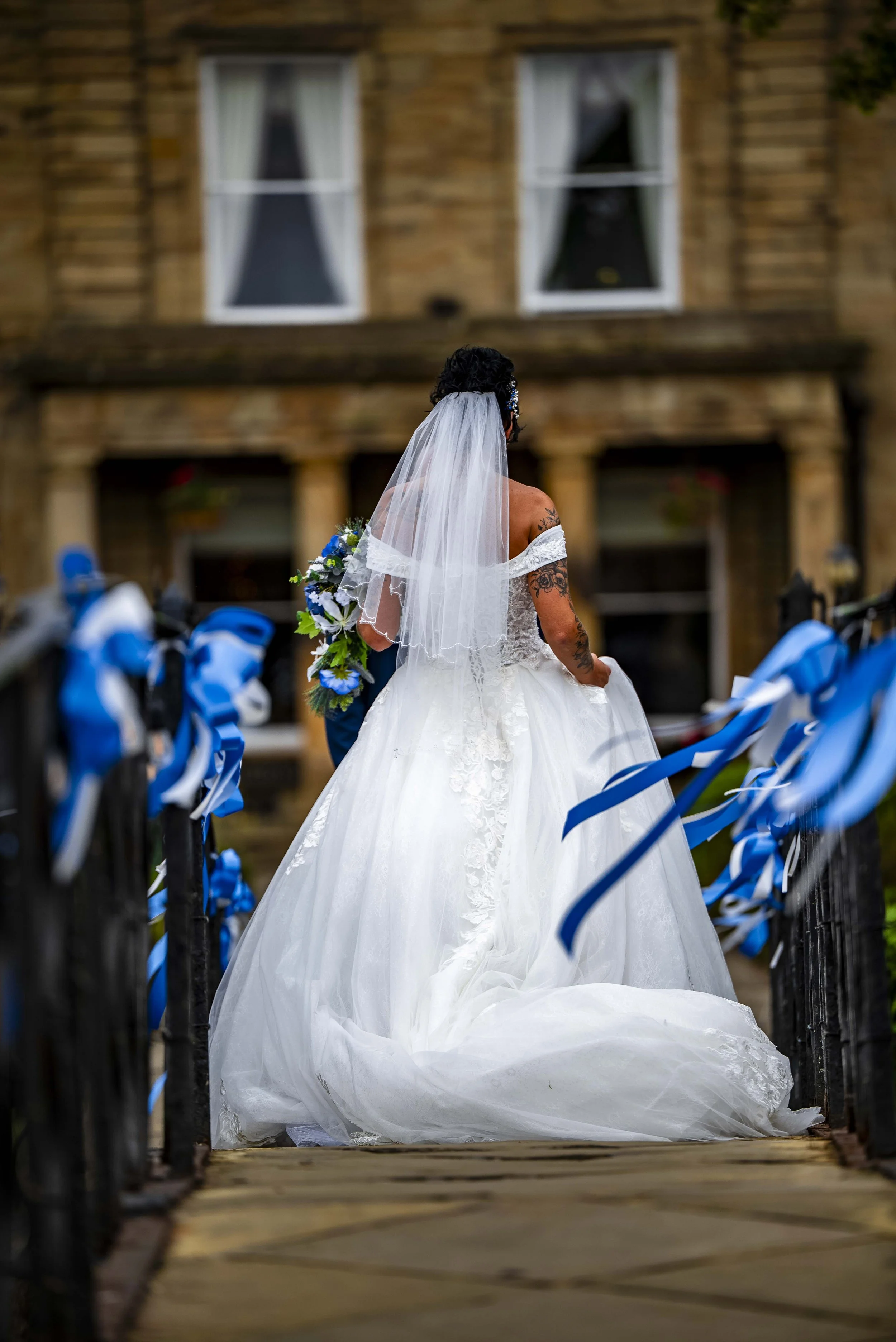 A bride in a white wedding gown and veil is standing on a bridge decorated with blue ribbons, holding a bouquet, with a building in the background.