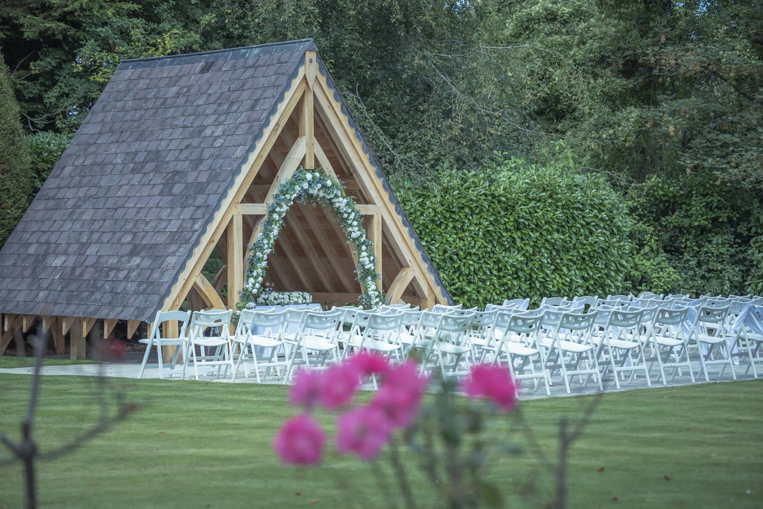 Outdoor wedding ceremony setup with white chairs facing a small wooden A-frame structure decorated with white flowers and greenery, surrounded by lush green trees and manicured grass.