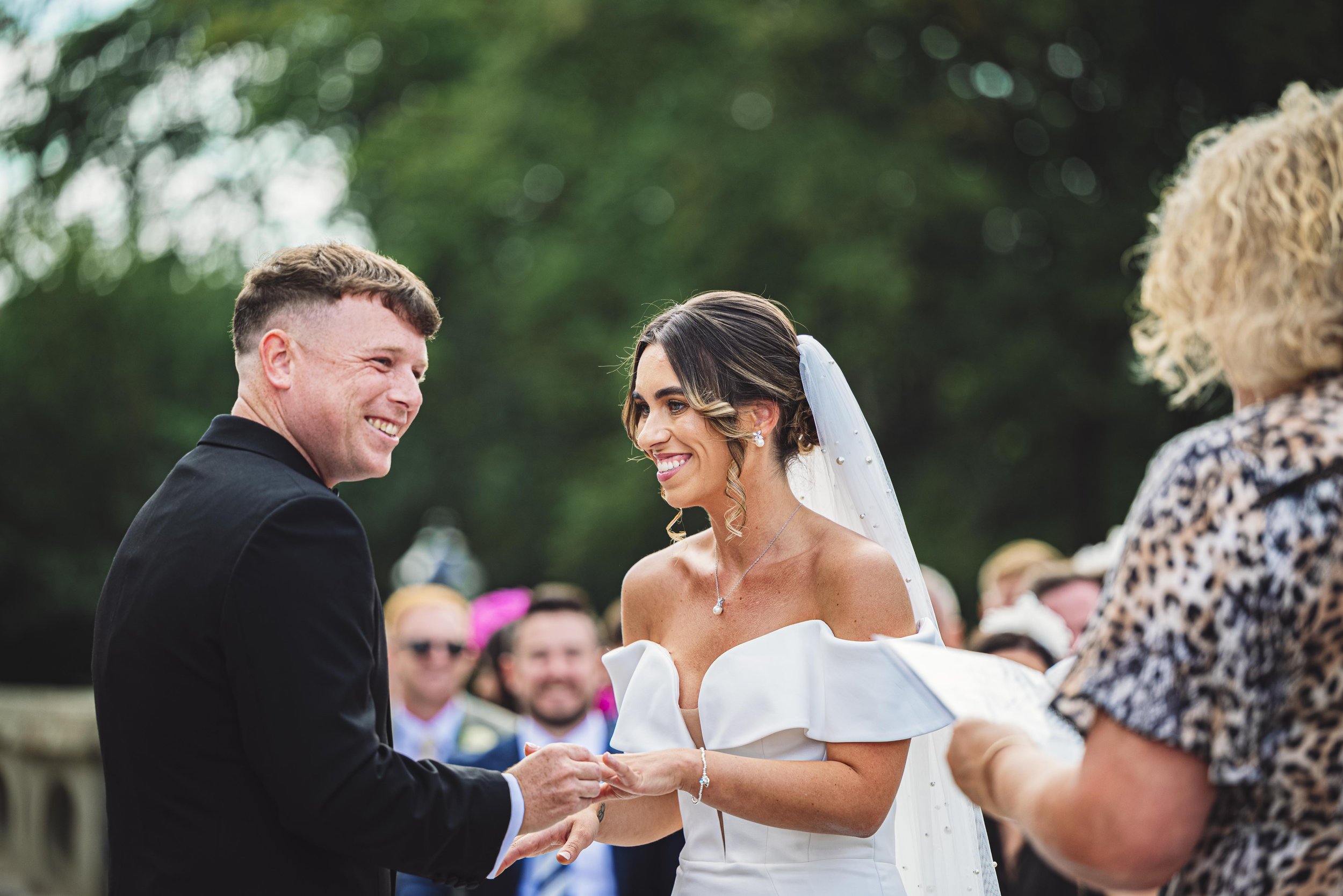 Bride and groom exchanging wedding rings outdoors, with guests watching.