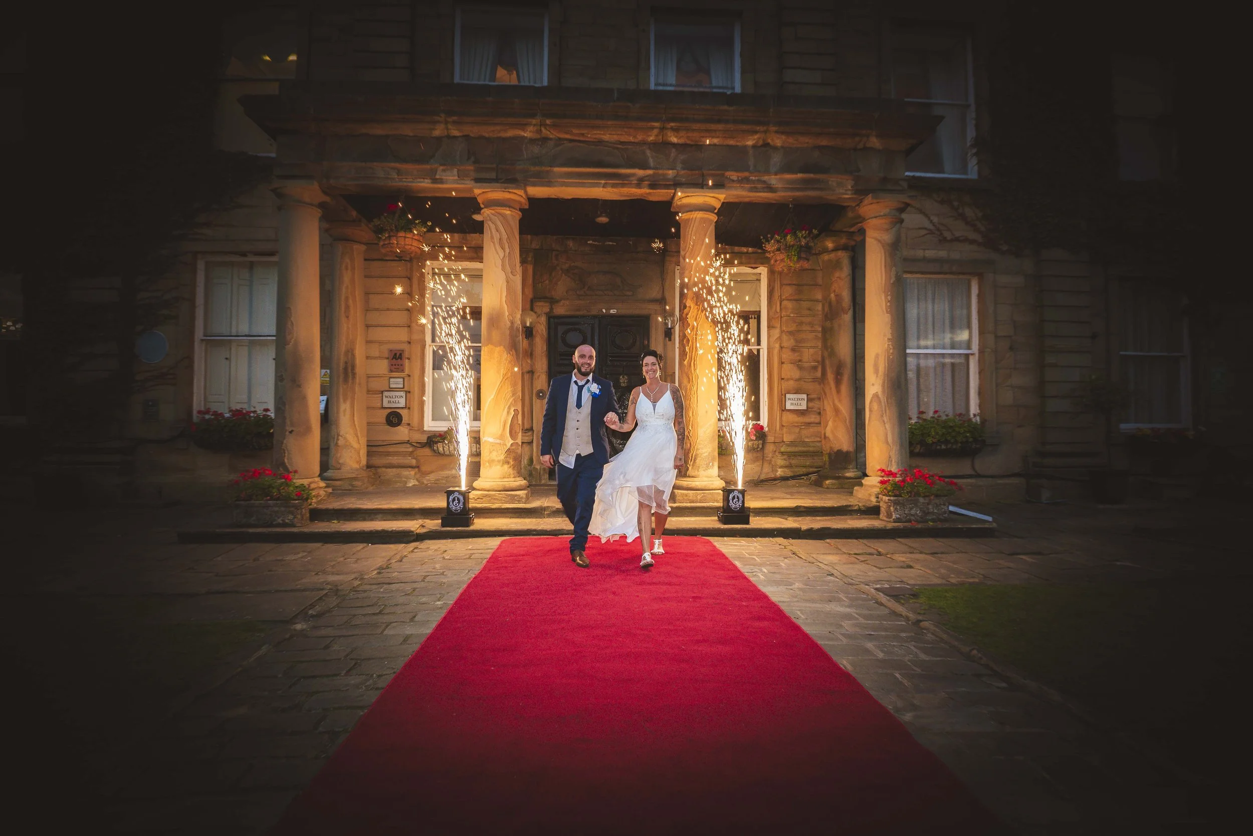 A newlywed couple walking on a red carpet outside a historic building with firework sparklers, flowers, and lanterns at night.