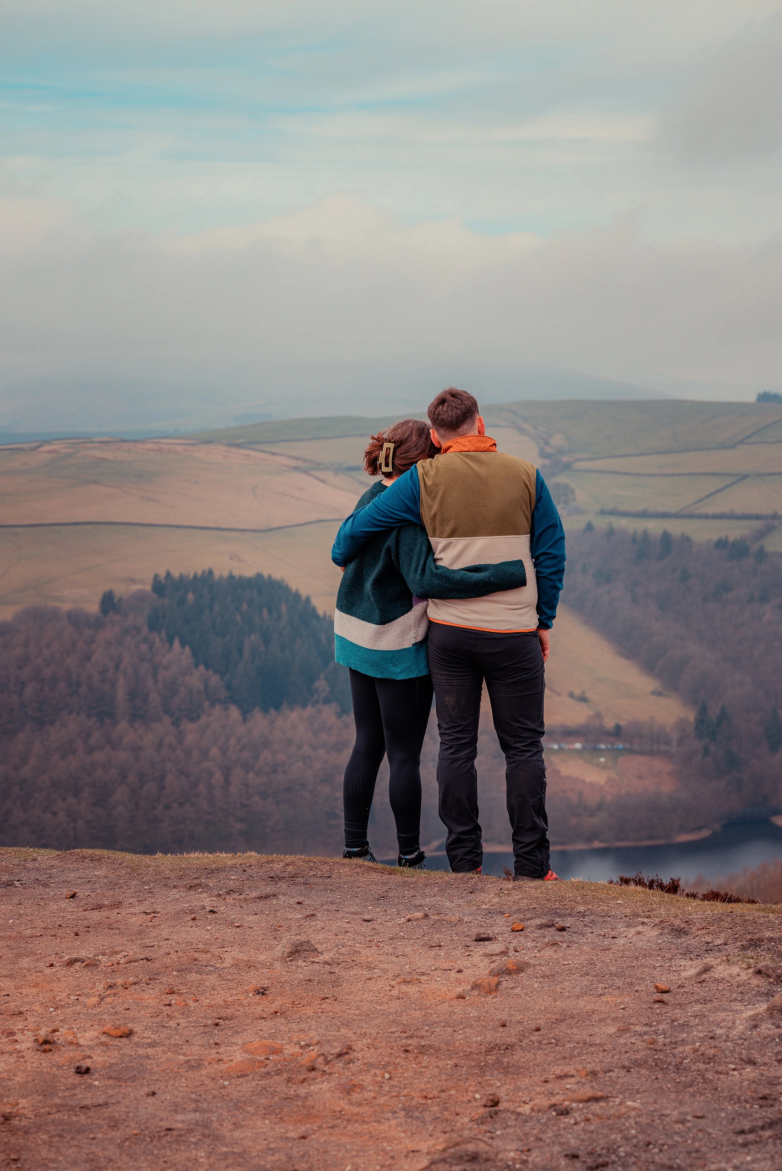 A couple standing on a hilltop, looking at a landscape of rolling hills and trees under an cloudy sky.