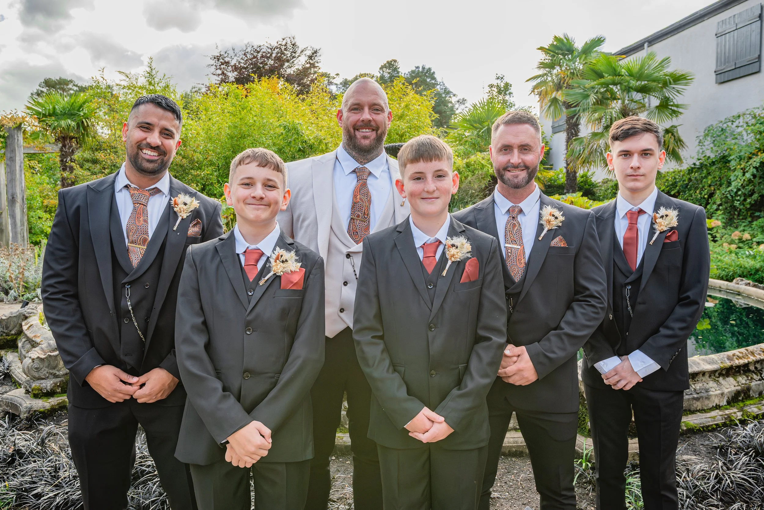 Group of seven men dressed in formal suits, smiling outdoors in a garden with trees and plants.
