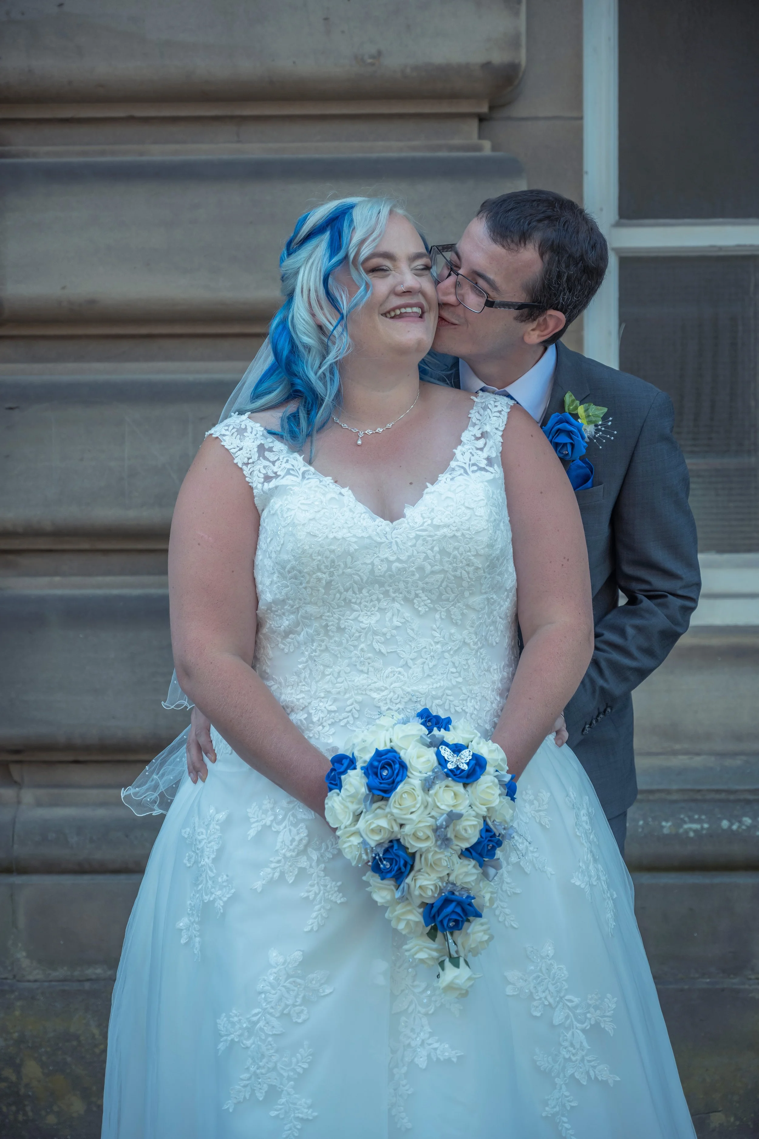 A bride with blue and white hair in a white wedding dress, holding a bouquet of white and blue roses, stands outside a building. A groom in a gray suit leans in to kiss her on the cheek.