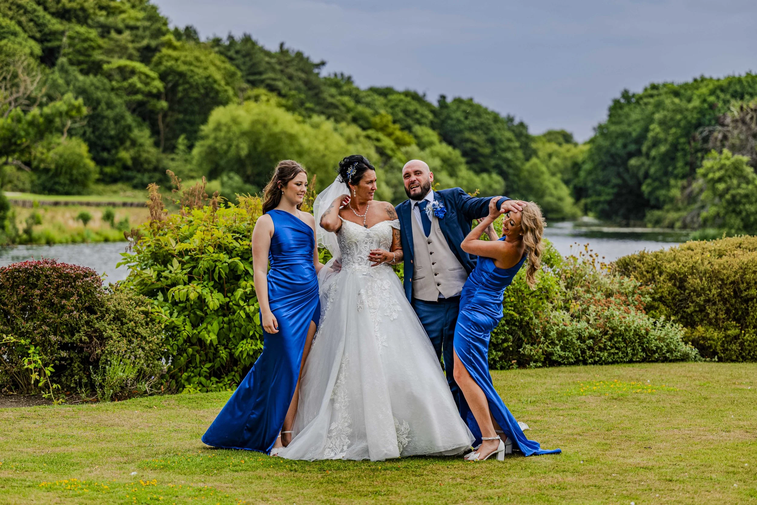 A wedding party outdoors with a bride in a white gown and three women in blue dresses, near a river and lush green trees.