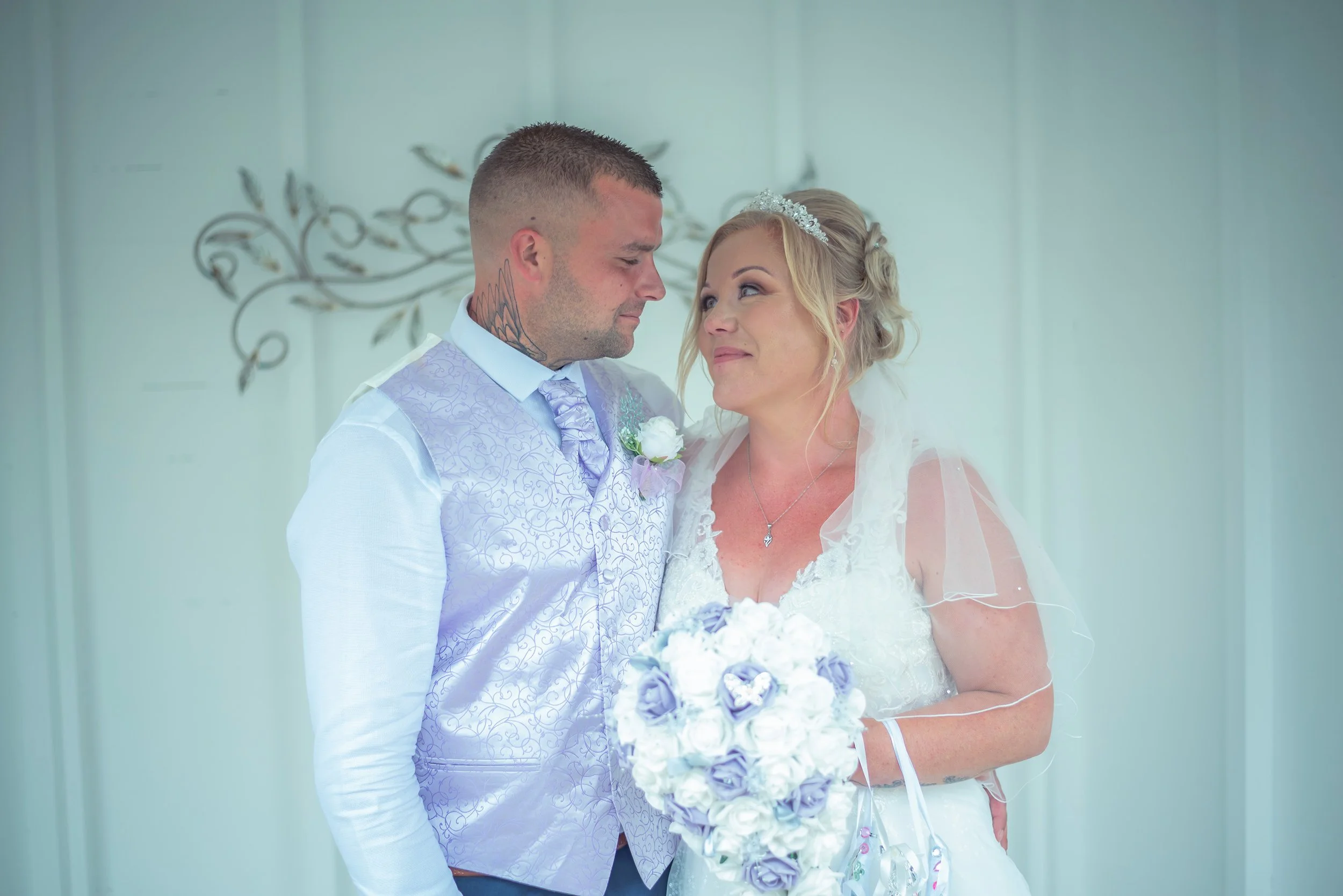 A bride and groom standing close together, looking into each other's eyes, during a wedding ceremony. The groom has a tattoo on his neck and is wearing a light purple vest and tie, while the bride has a tiara and is holding a bouquet of white and purple flowers.