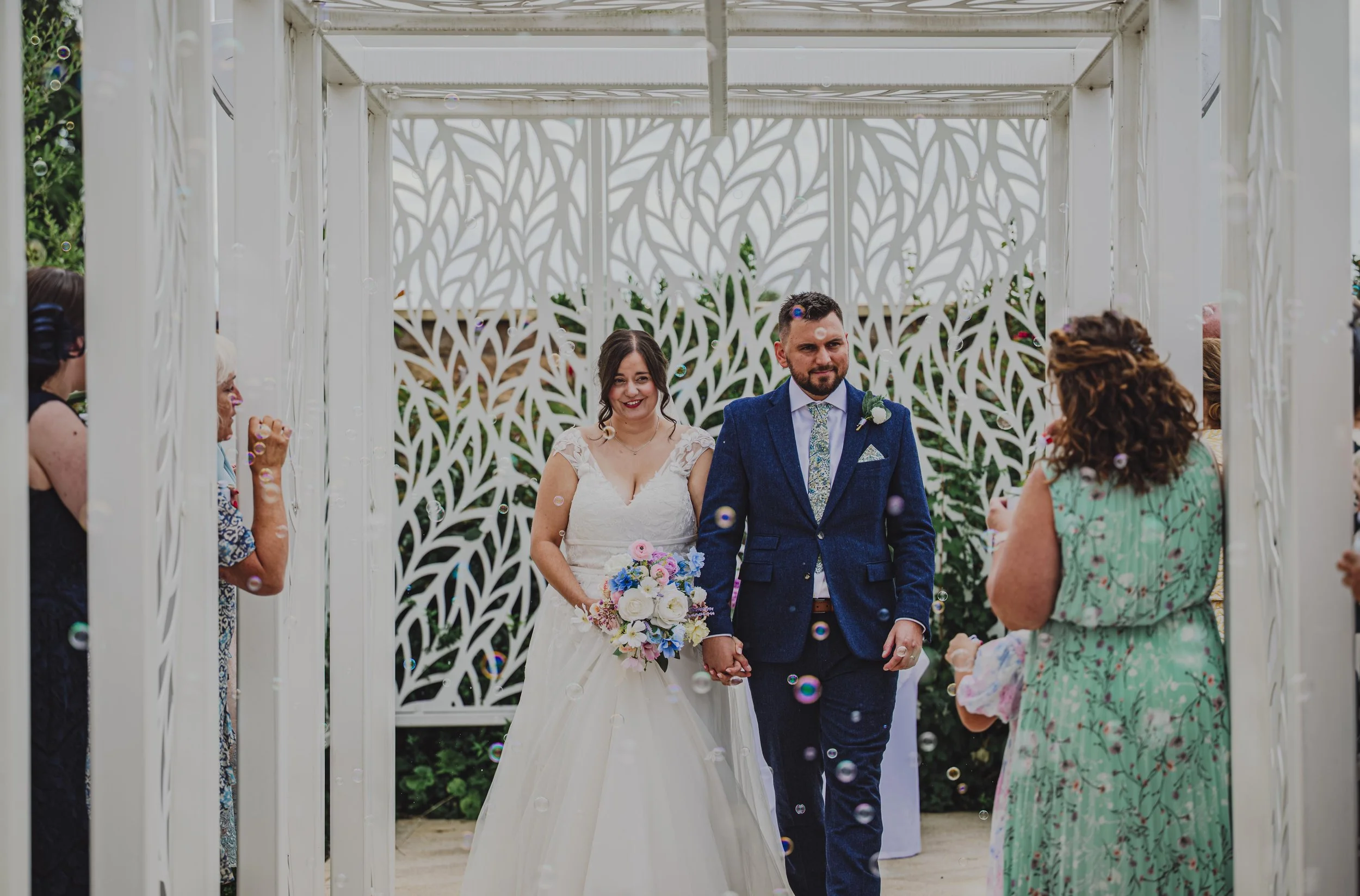 A bride and groom walking hand in hand under a decorated white pergola after their wedding ceremony, surrounded by friends and family, with bubbles floating around.
