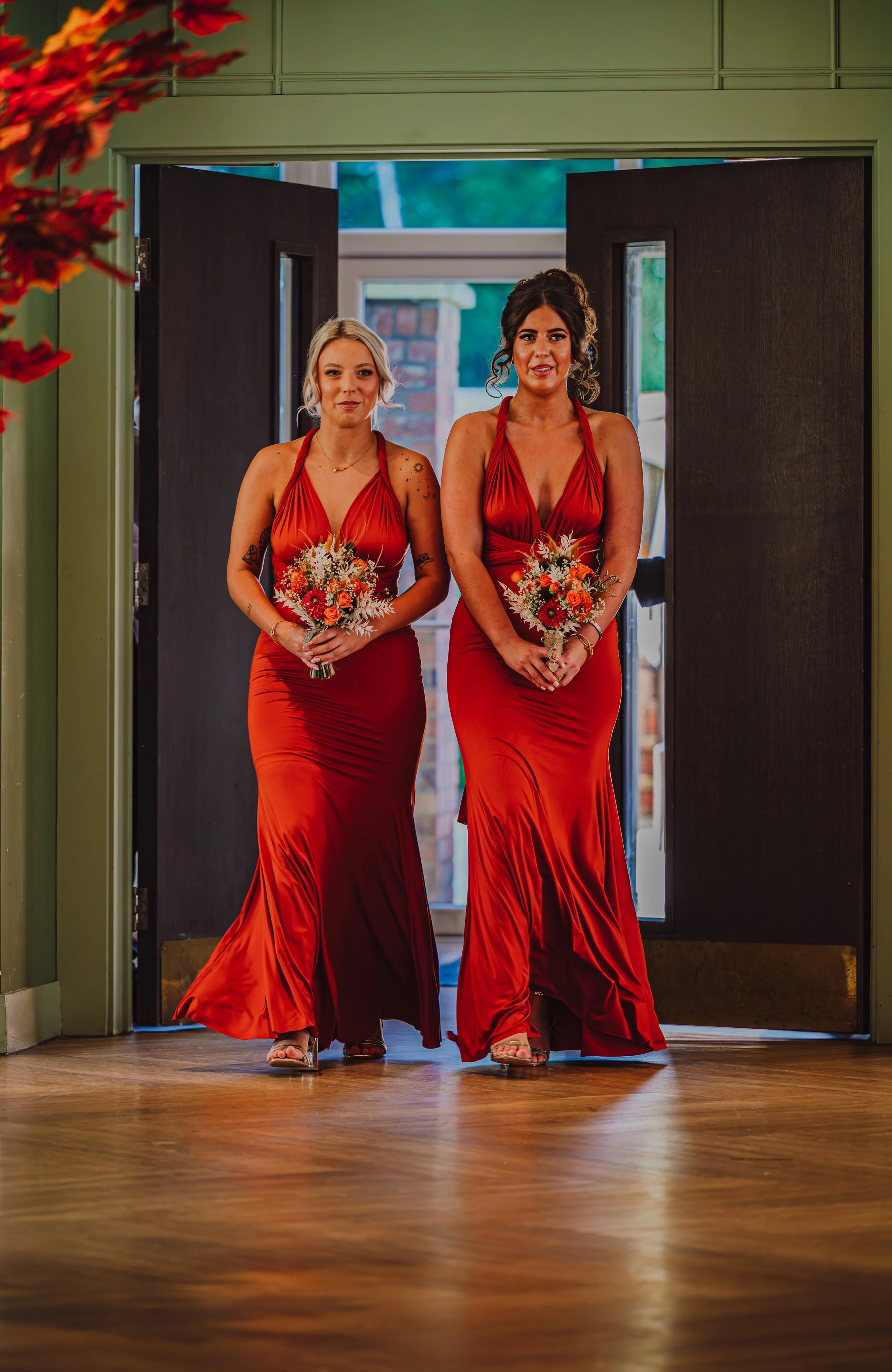 Two women in matching red dresses holding bouquets walk through a doorway into a room with wooden floors. One woman has blonde hair, the other dark curly hair. They are dressed for a formal occasion.