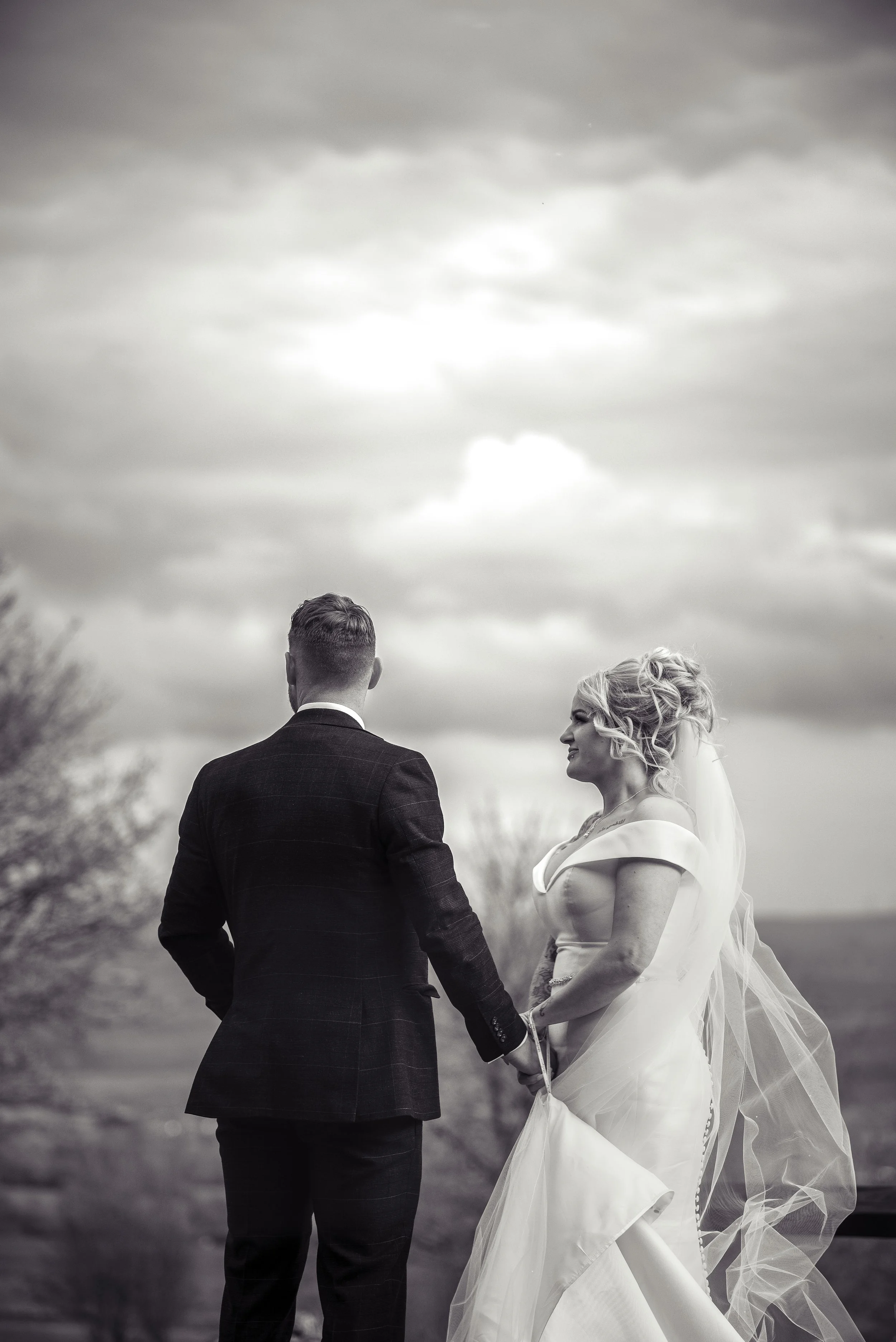 A black and white photo of a bride and groom holding hands outdoors, facing each other with a cloudy sky in the background.
