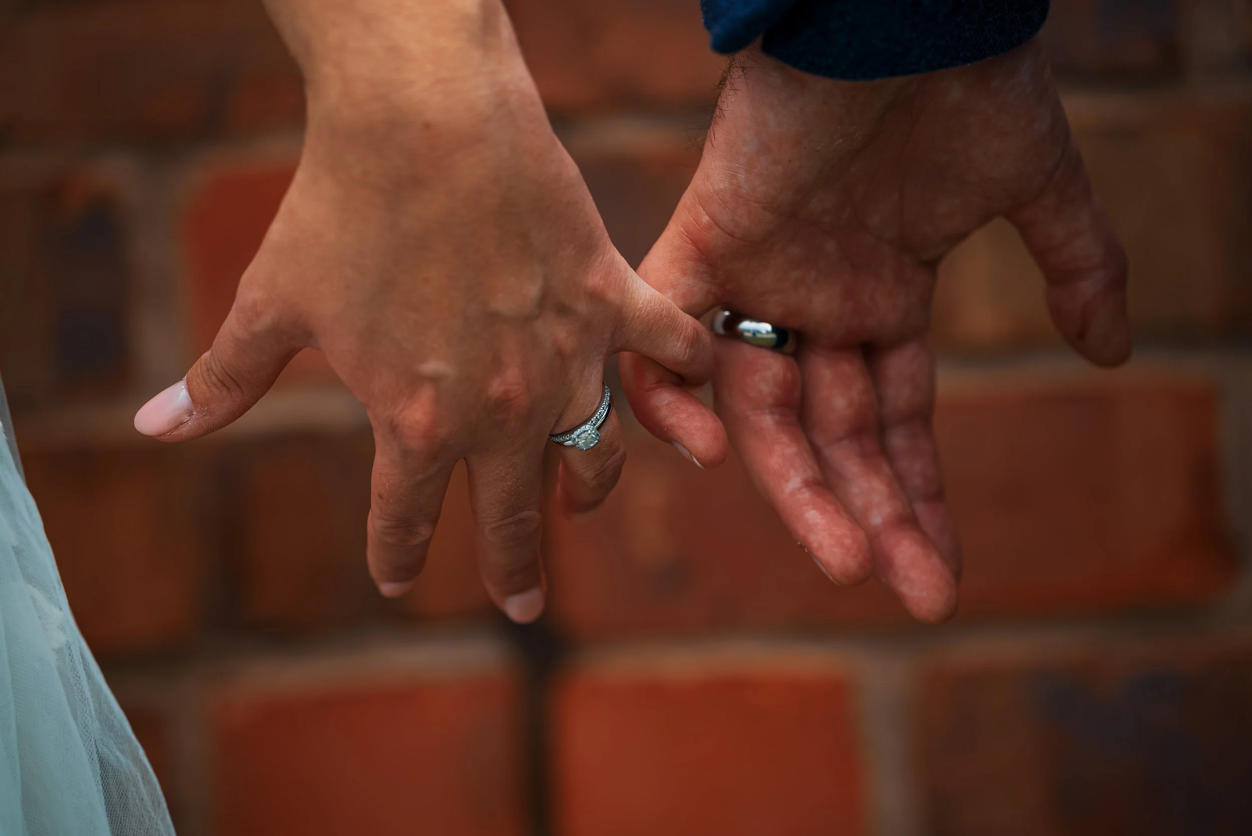 Close-up of a man and woman holding hands, with the woman's hand featuring a diamond engagement ring. The background shows a blurred brick wall.