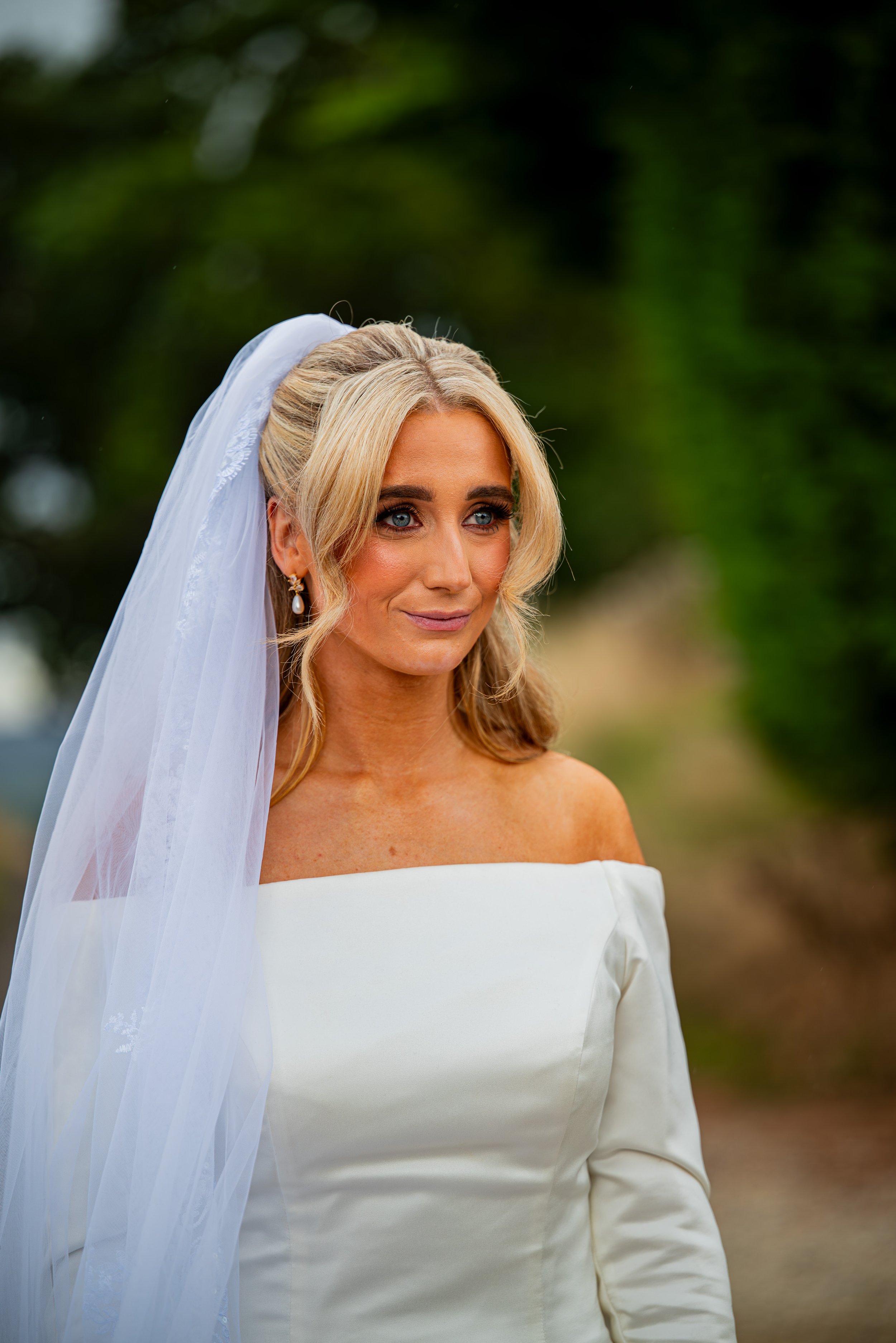 A bride in a white off-the-shoulder wedding dress and veil standing outdoors with a blurred green background.