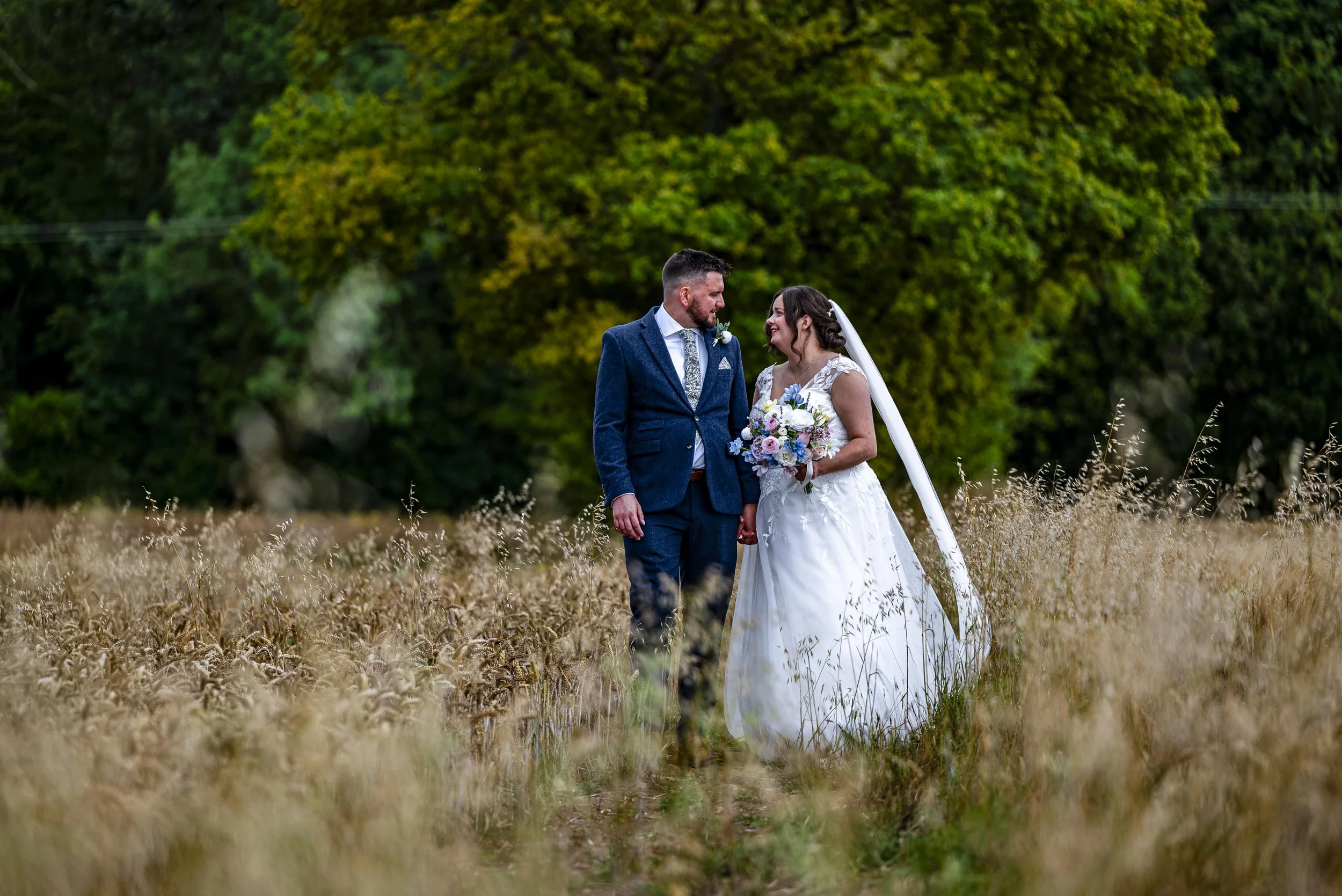 A bride and groom standing in a field of tall grass, holding hands and smiling at each other, with trees in the background.