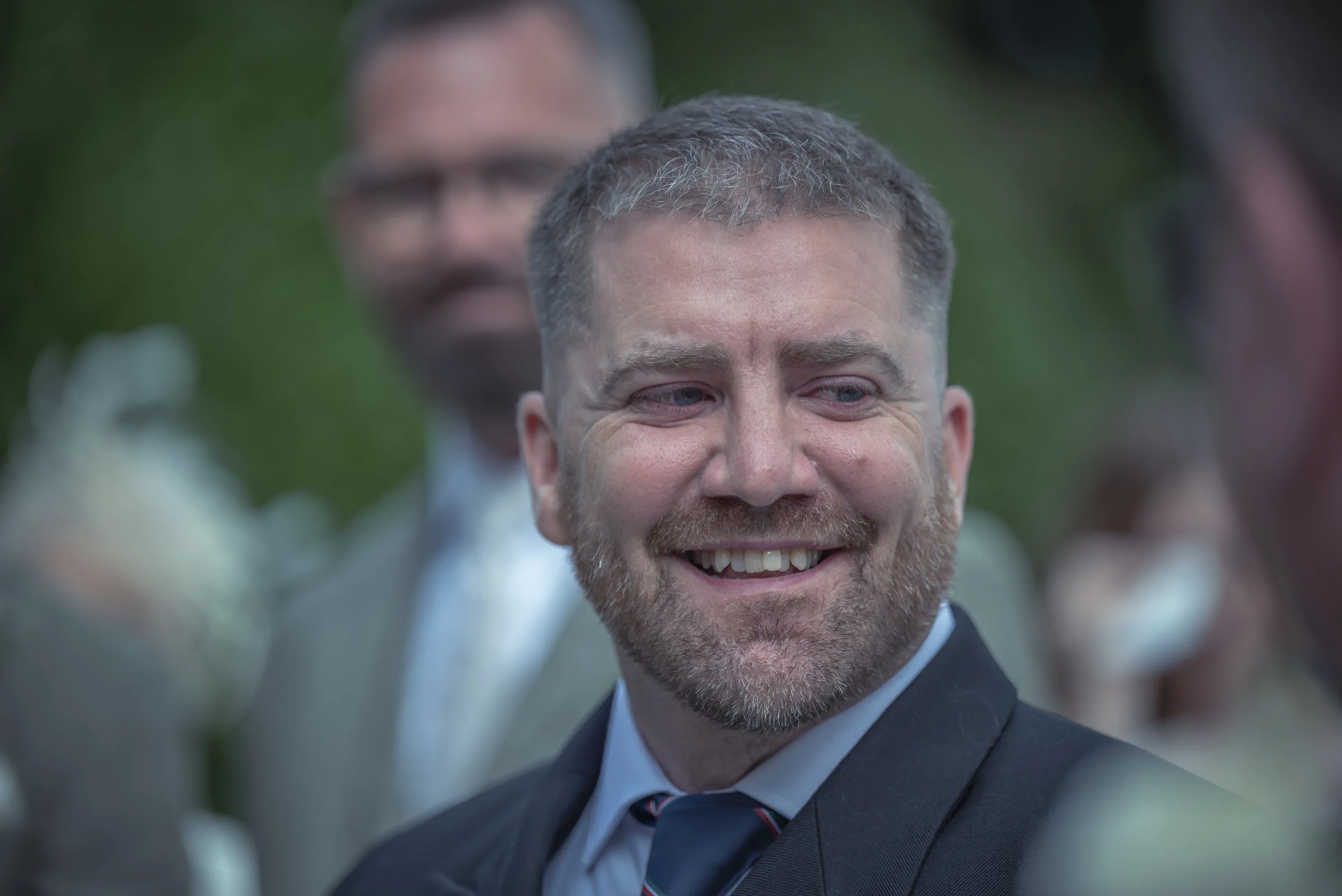 A smiling man with a beard in a suit at an outdoor event, with blurred people in the background.