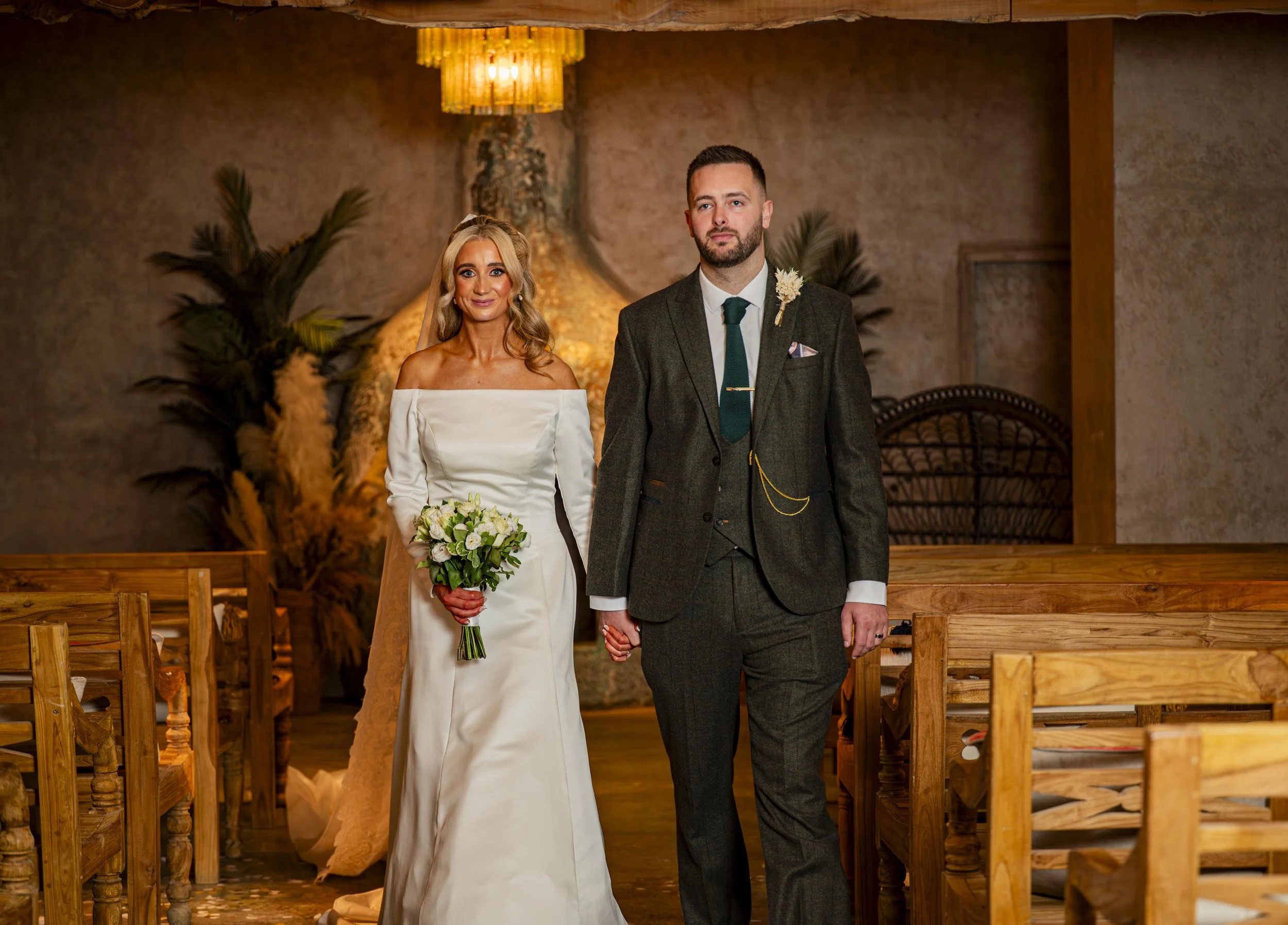 A bride and groom standing hand in hand inside a rustic wedding venue with wooden pews and decorative plants, with warm lighting and a chandelier overhead.