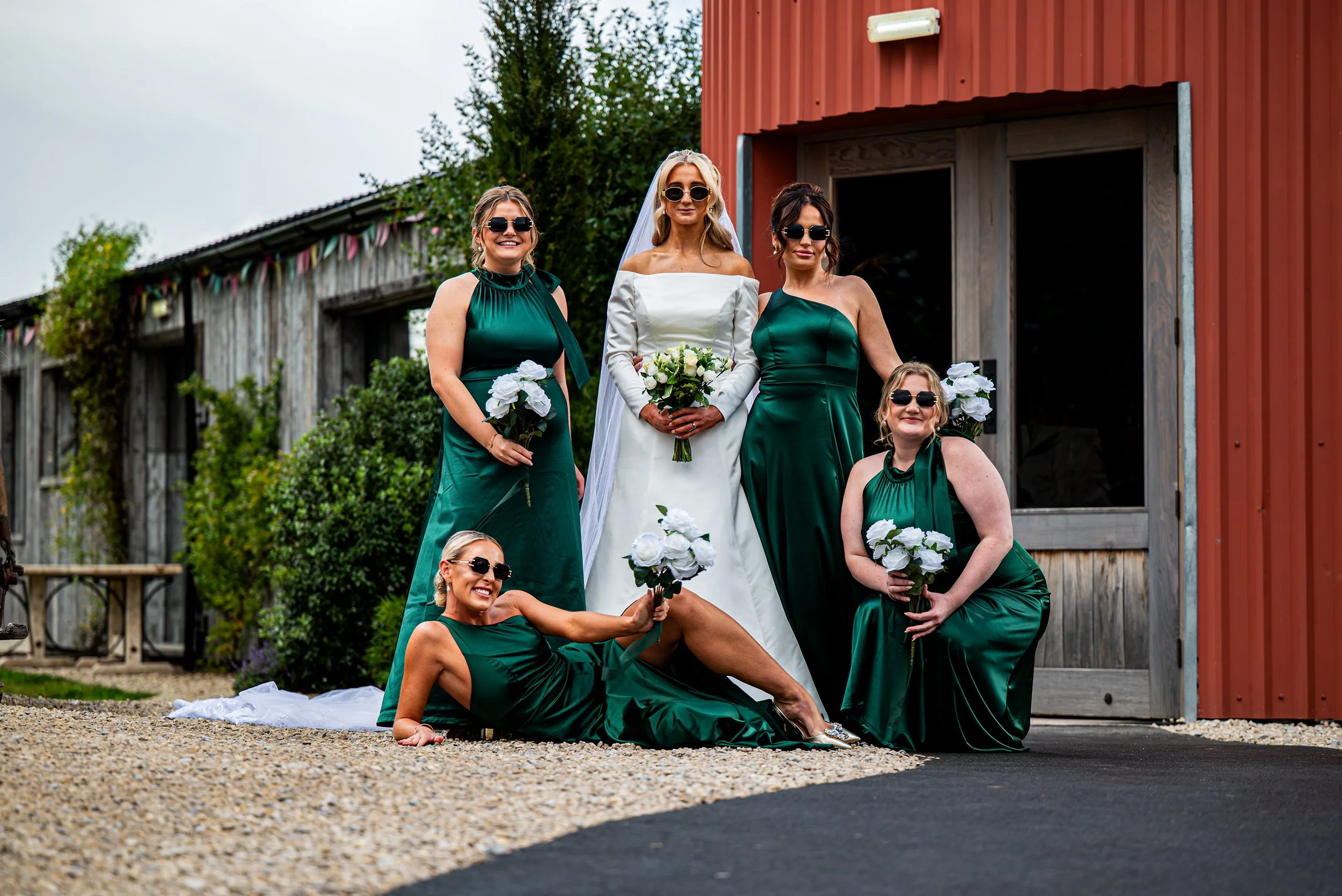 Bride and five bridesmaids in green dresses with white bouquets, posing outside in front of rustic barns on a cloudy day.