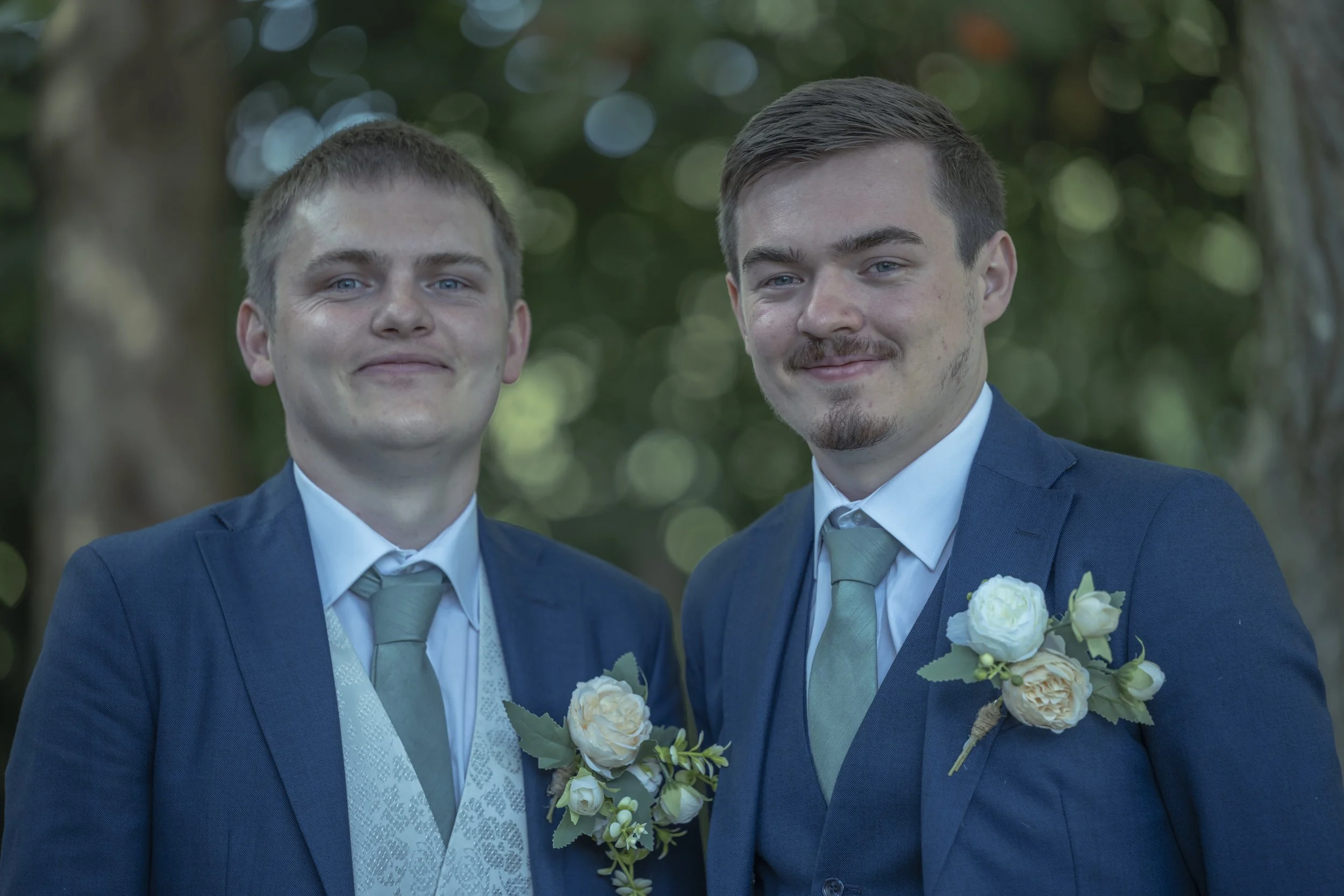 Two men in blue suits with matching green ties and boutonnières on lapels standing outdoors with a wooded background, smiling at the camera.
