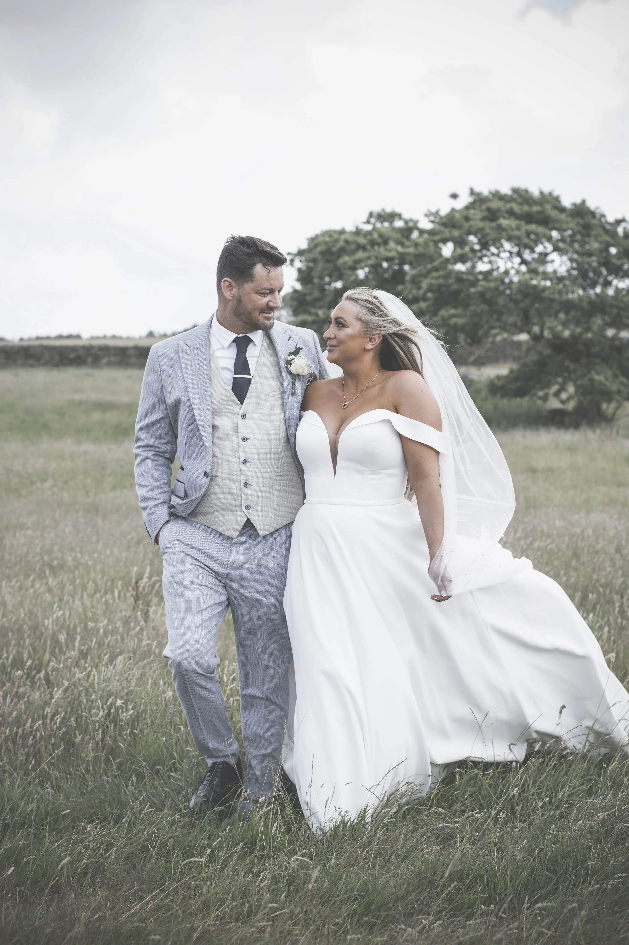 A newlywed couple standing in a field outdoors, looking at each other. The man is wearing a light gray suit and tie, and the woman is wearing a white wedding dress with off-the-shoulder sleeves and a veil.