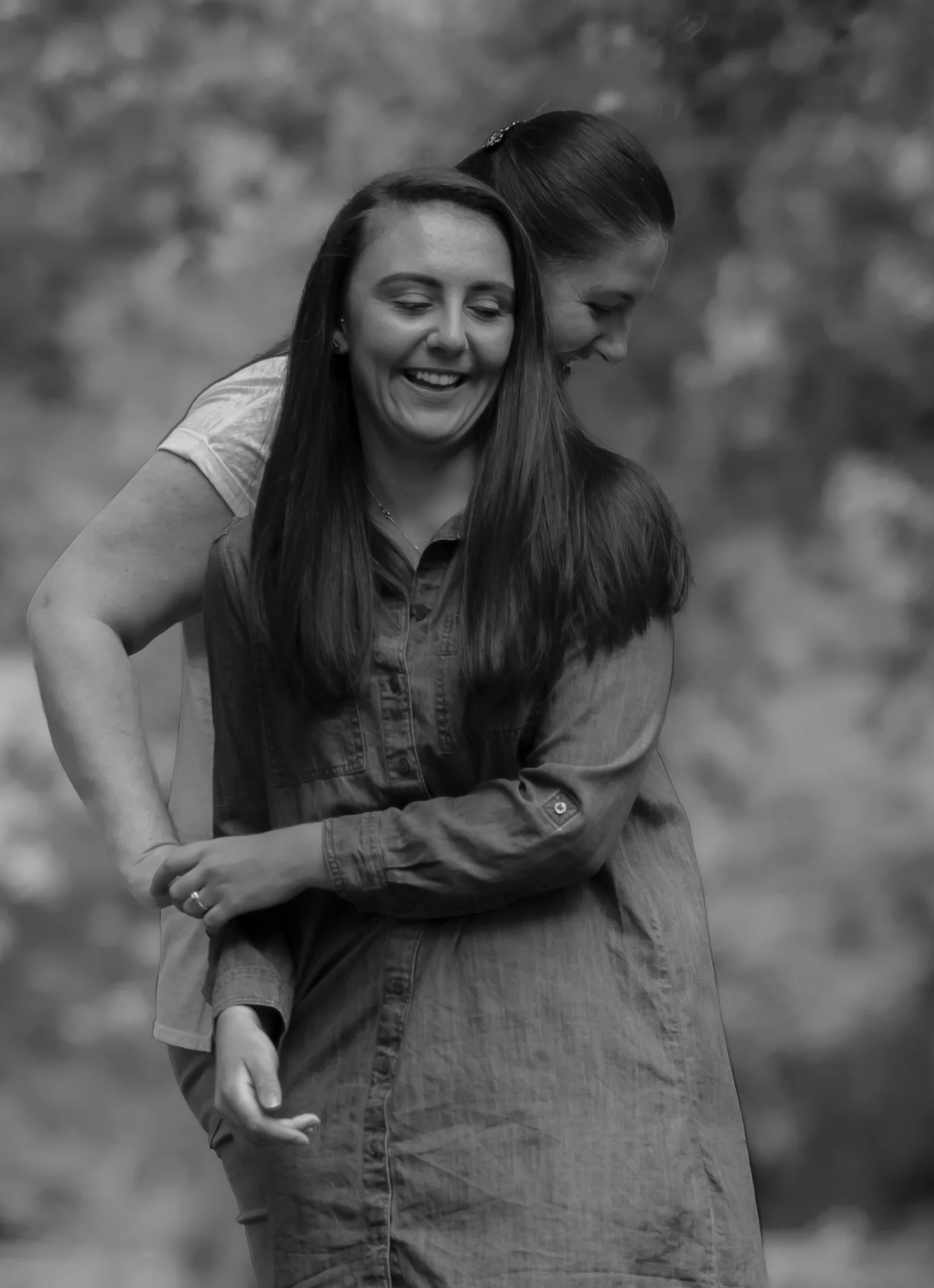 Two women smiling and embracing outdoors, one with long dark hair and the other with them all busy