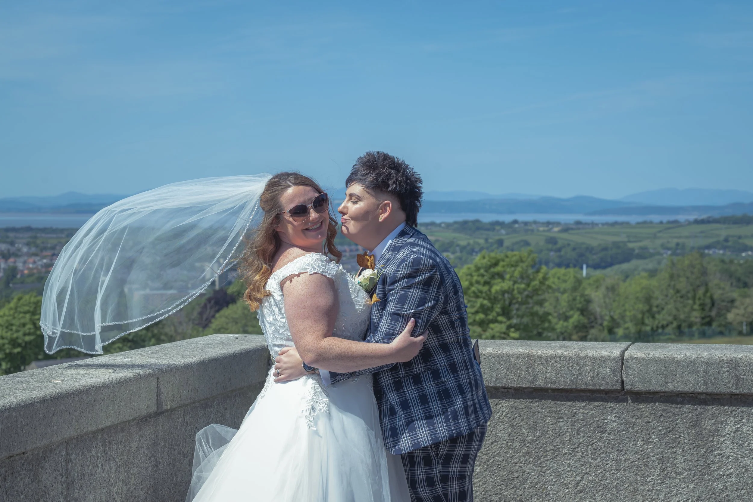 A bride and groom sharing an embrace outdoors on a sunny day, with a scenic view of green trees, water, and mountains in the background.