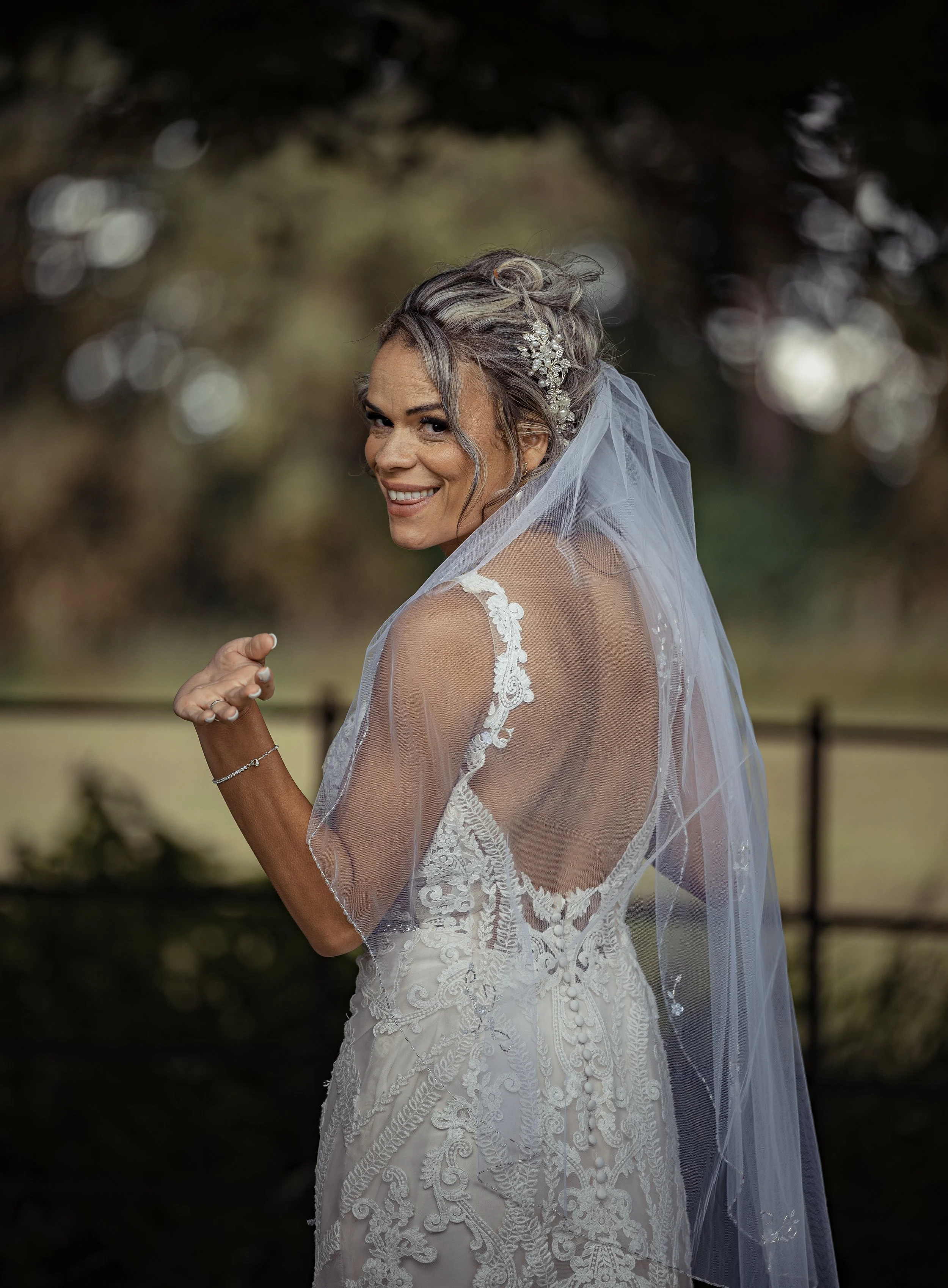 A smiling bride in a wedding dress with lace details and a veil is outdoors, with blurred trees in the background.