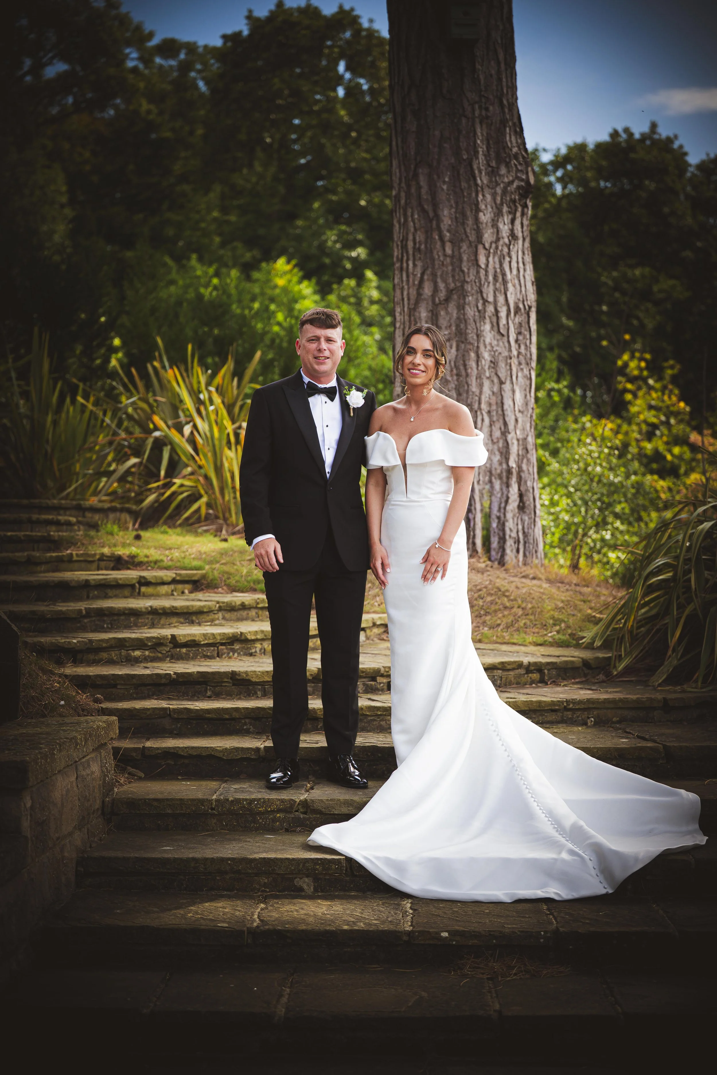 A bride and groom in wedding attire standing on stone stairs outdoors with greenery and a large tree in the background.