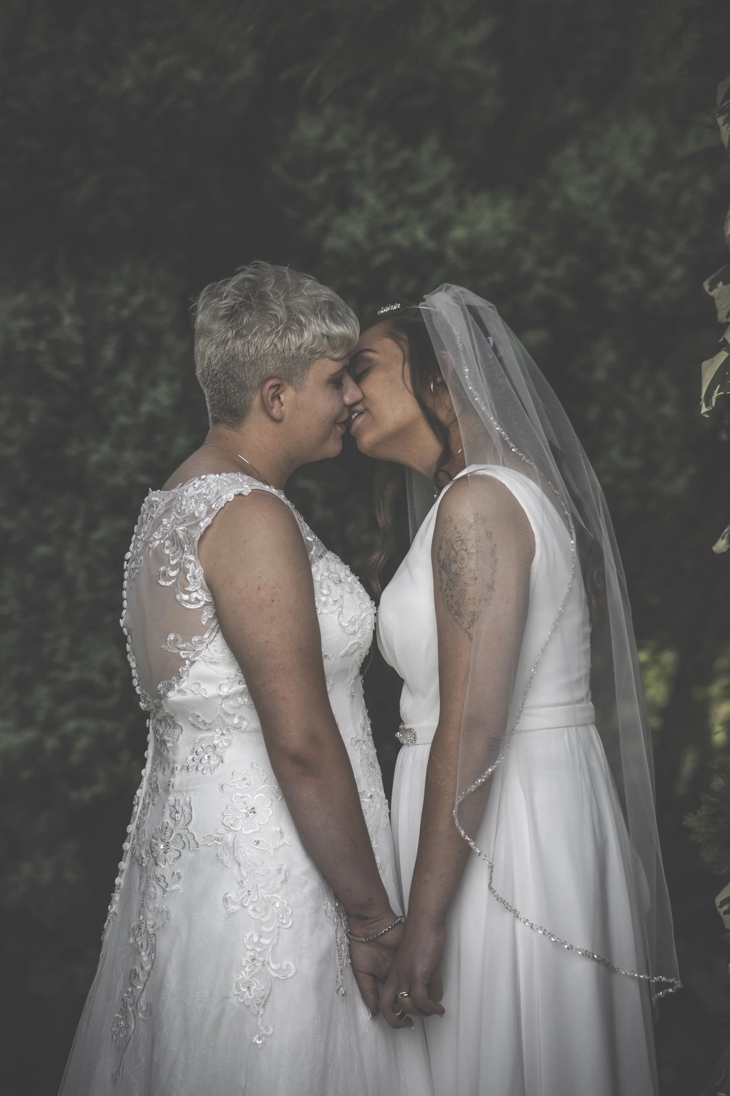 Two women in wedding dresses touching foreheads and holding hands outdoors, with a dark green leafy background.