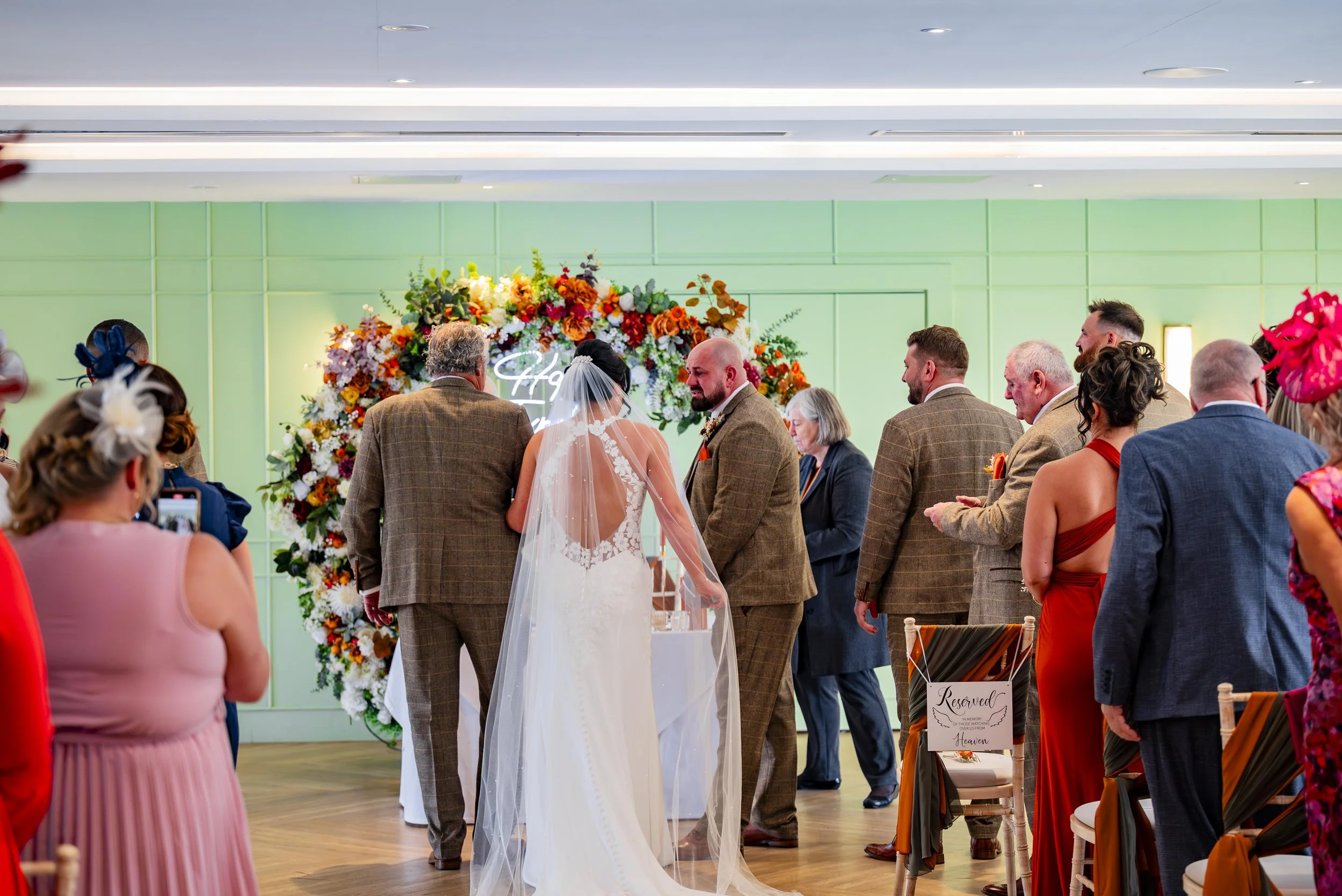 A wedding ceremony with the bride and groom standing in front of a floral arch, surrounded by guests. The bride wears a white dress with a veil, and the groom wears a brown plaid suit. The background features a green wall with a neon sign that reads 