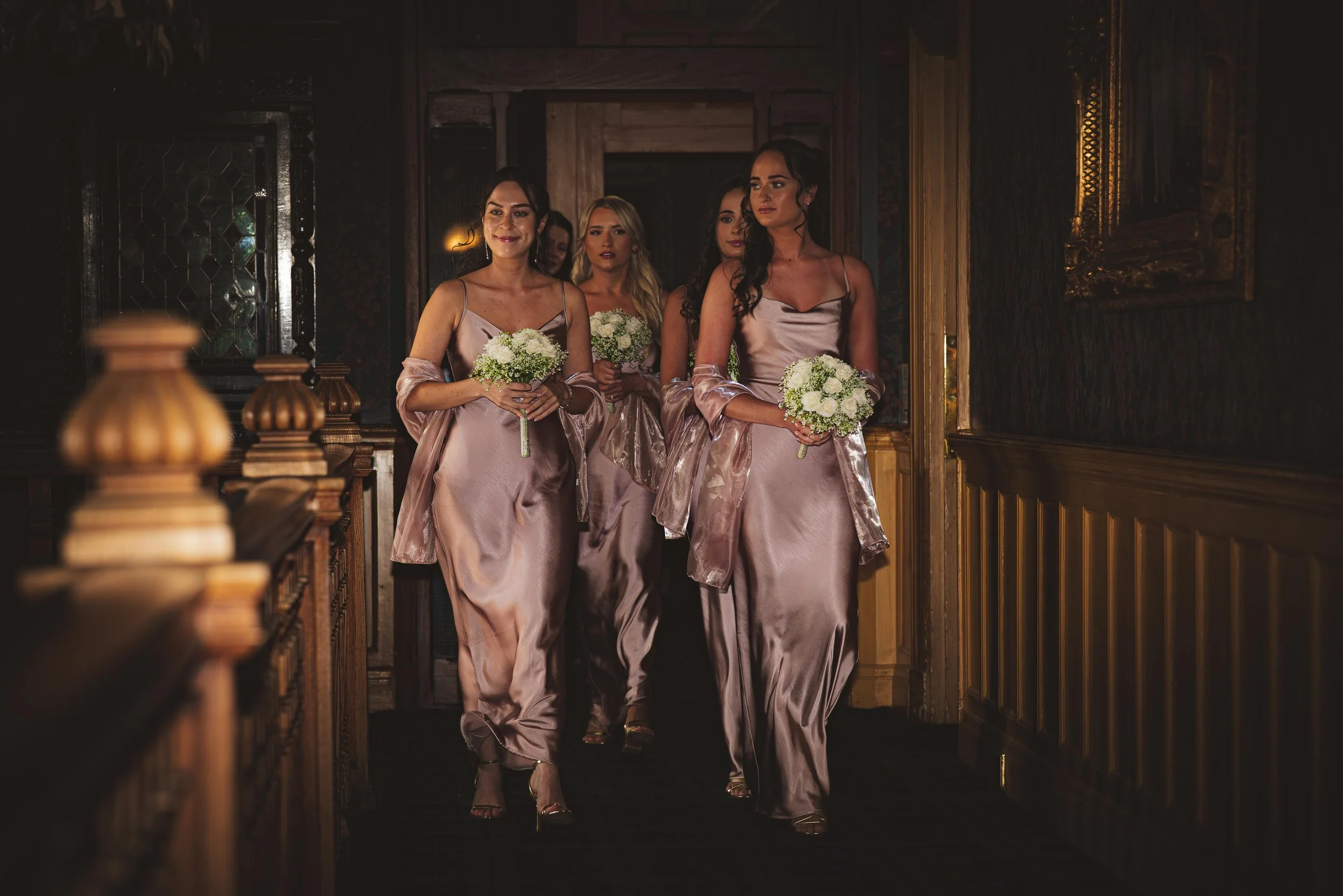 Bridesmaids in mauve satin dresses walking down a wooden corridor holding white floral bouquets.