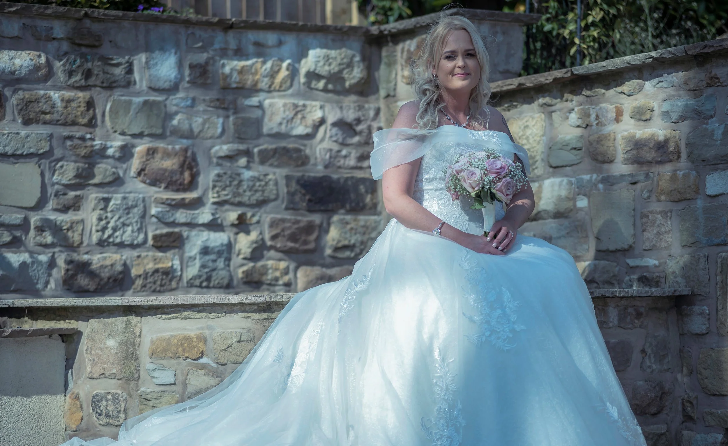 A woman in a white wedding dress holding a bouquet of pink roses, sitting on a stone bench in front of a stone wall.