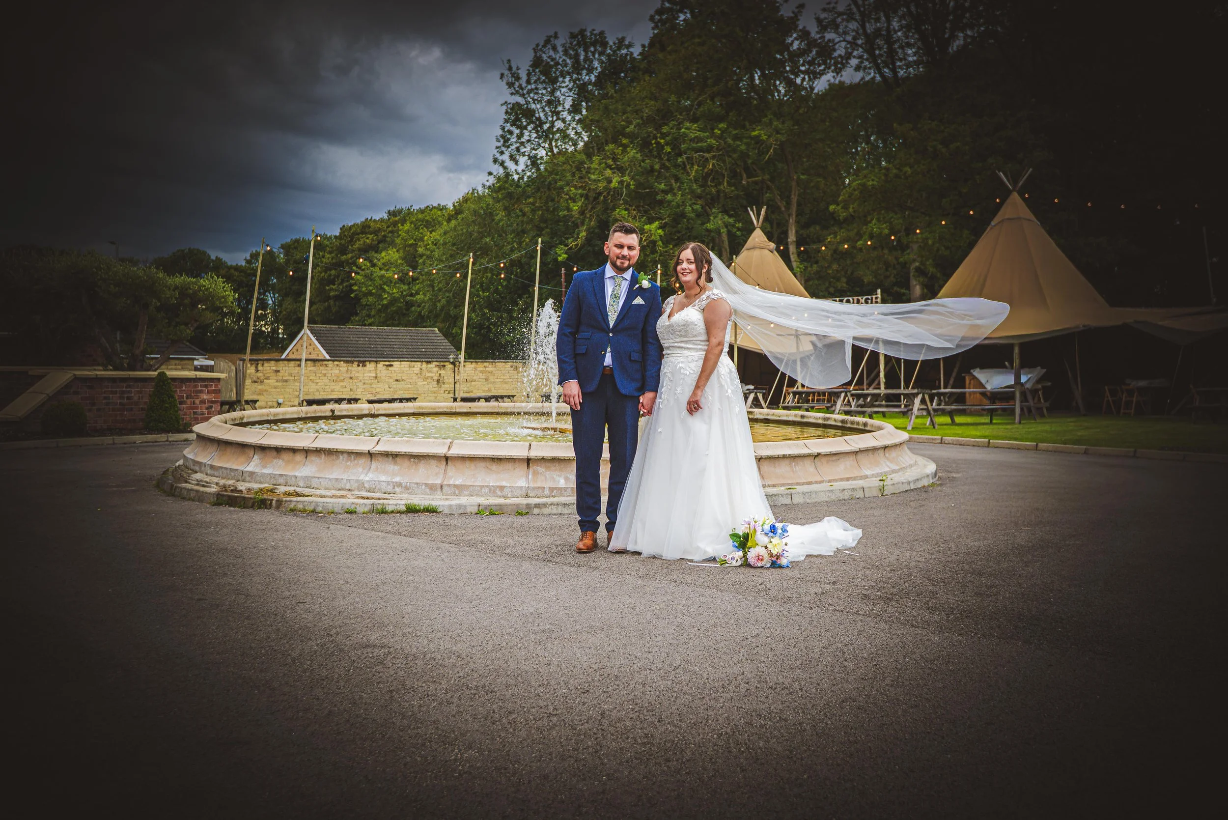 Bride and groom standing together outdoors in front of a fountain during cloudy weather at a wedding.