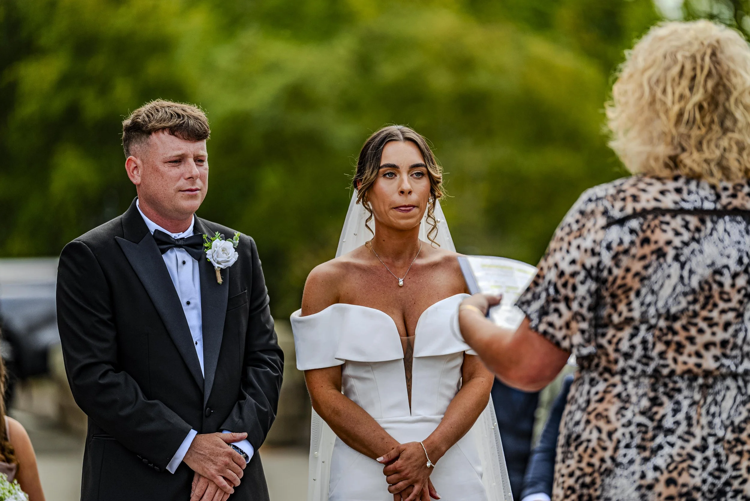 A bride and groom standing outdoors during a wedding ceremony, facing a woman holding papers, with a green background.
