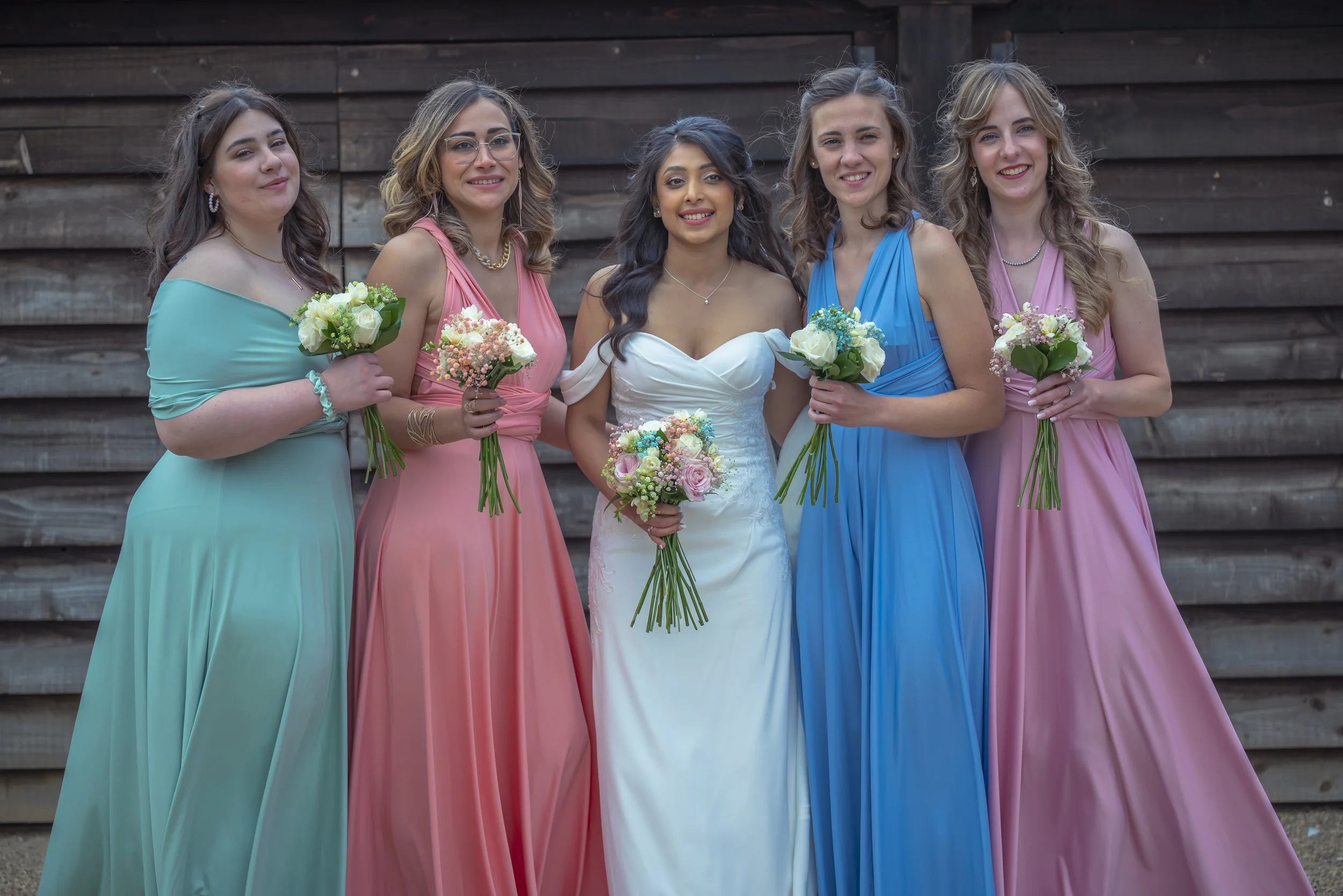Six women dressed in colorful gowns, standing together and holding bouquets of flowers, in front of a wooden wall.
