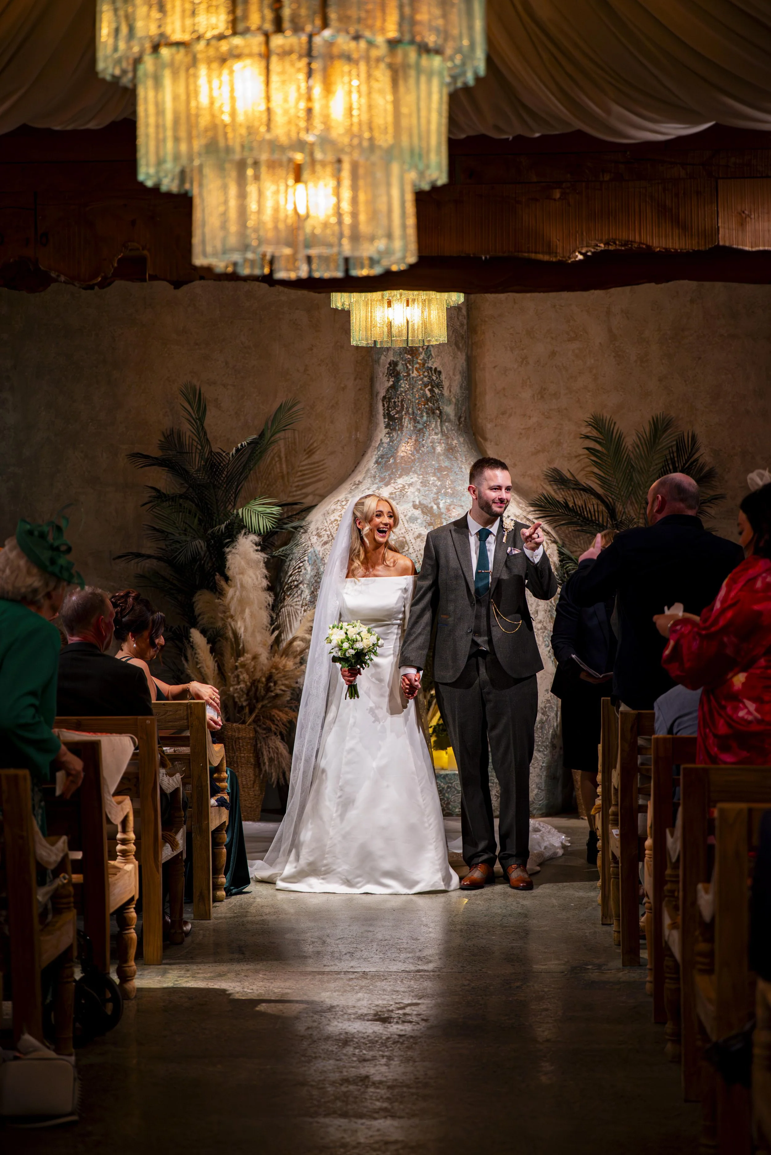 A bride and groom holding hands during their wedding ceremony, standing before an officiant, with guests seated on either side, in a warmly decorated indoor venue with large chandeliers and greenery.