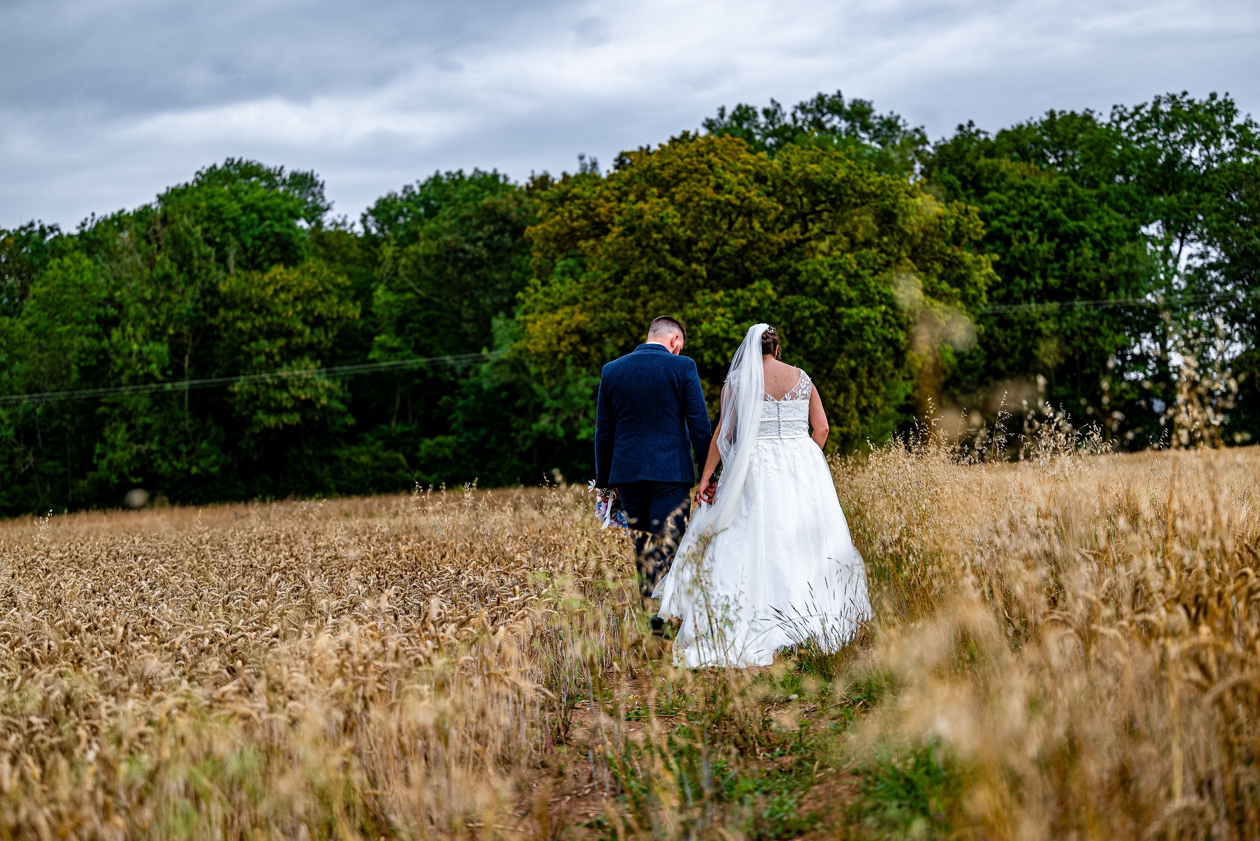 Bride and groom walking through a wheat field, holding hands, with green trees and a cloudy sky in the background.