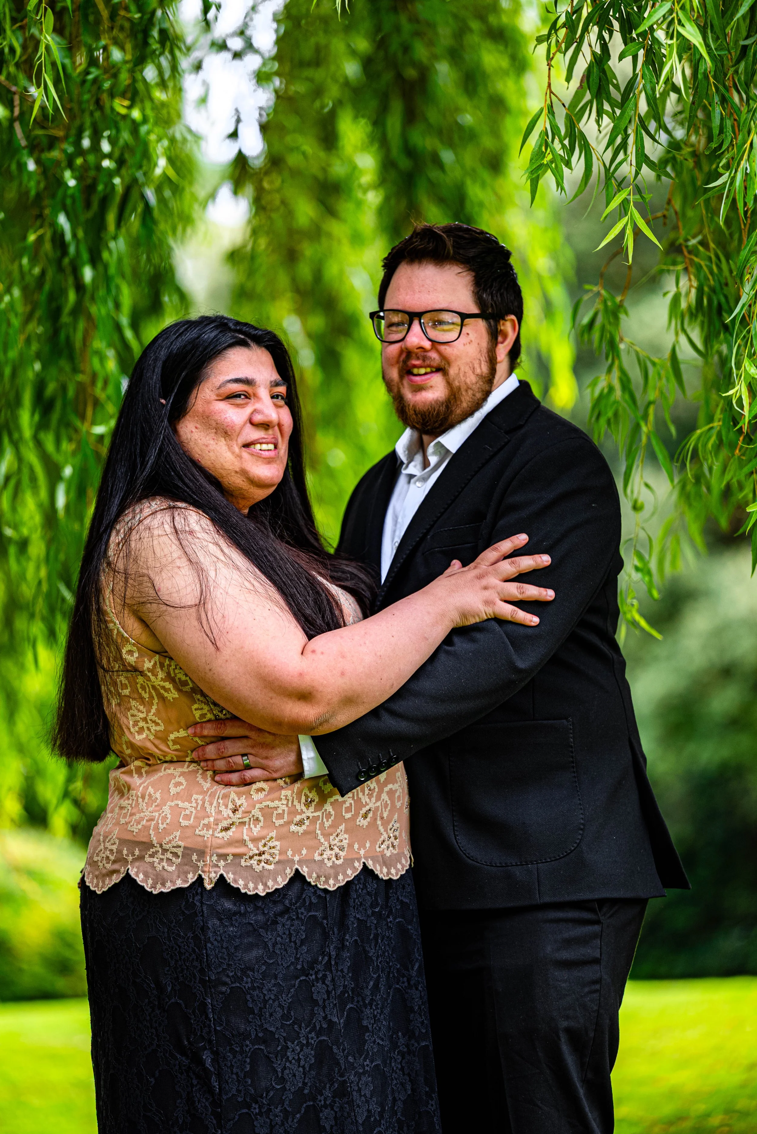 A couple standing close together outdoors, surrounded by green trees and foliage, smiling for a photograph.